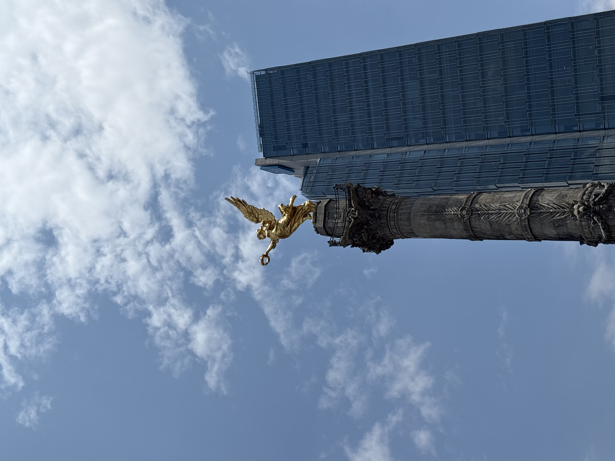 The image shows the Angel of Independence monument in Mexico City, Mexico. The golden statue of the winged Victory is perched atop a tall, ornate column. A modern, glass-clad skyscraper rises behind the monument. The sky is blue with scattered white clouds.
