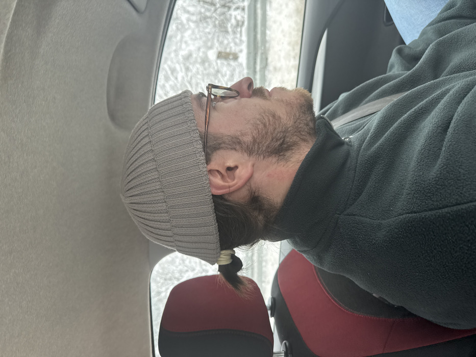 An adult male is depicted in the front passenger or driver's seat of a vehicle in Tutin Municipality, Serbia. He is observed in right profile, wearing a light gray ribbed beanie hat, eyeglasses with light brown frames, and a dark green fleece jacket. His hair, secured in a small ponytail with a light-colored elastic at the nape of his neck, is visible beneath the beanie. A beard and mustache cover his lower face. A diagonal seatbelt crosses his chest. To his left, a car seat with dark red and black upholstery is visible. The vehicle's interior features a light-colored headliner. Through the windows, a blurred, snow-covered outdoor environment is apparent, suggesting winter conditions. The man is stationary within the moving or parked vehicle, looking forward.