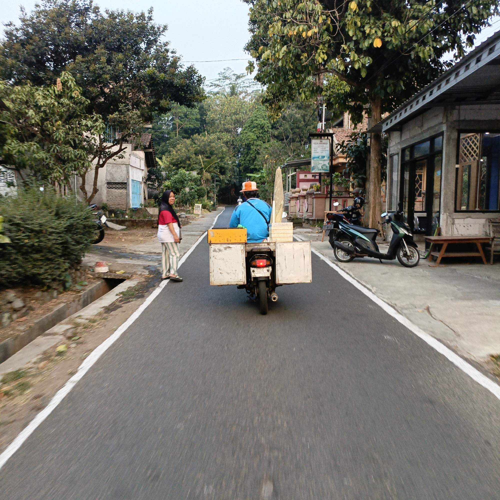 The image depicts a street scene in Salatiga, Indonesia, with a focus on a person riding a motorcycle equipped with a cargo carrier. The motorcycle, a dark color, is moving away from the camera on a paved road with a white line marking the edge. The rider is wearing a blue shirt and an orange and white hat, and is seated on the motorcycle. The cargo carrier is a rectangular box structure mounted behind the rider, with white side panels and a yellow top compartment.
To the left of the road, a woman wearing a headscarf, red shirt, and striped pants stands on a sidewalk. She is looking towards the motorcycle. Behind her, there is dense green foliage and a portion of a building. Further back, trees and other structures are visible.
To the right of the road, a building with a corrugated metal roof and a gray facade is present. There are windows on the building, and a wooden bench is placed in front of it. A dark green scooter is parked on the concrete area in front of the building. More trees and vegetation are seen in the background. The overall environment suggests a semi-urban or suburban setting with elements of nature integrated into the built environment. The lighting indicates it is daytime.