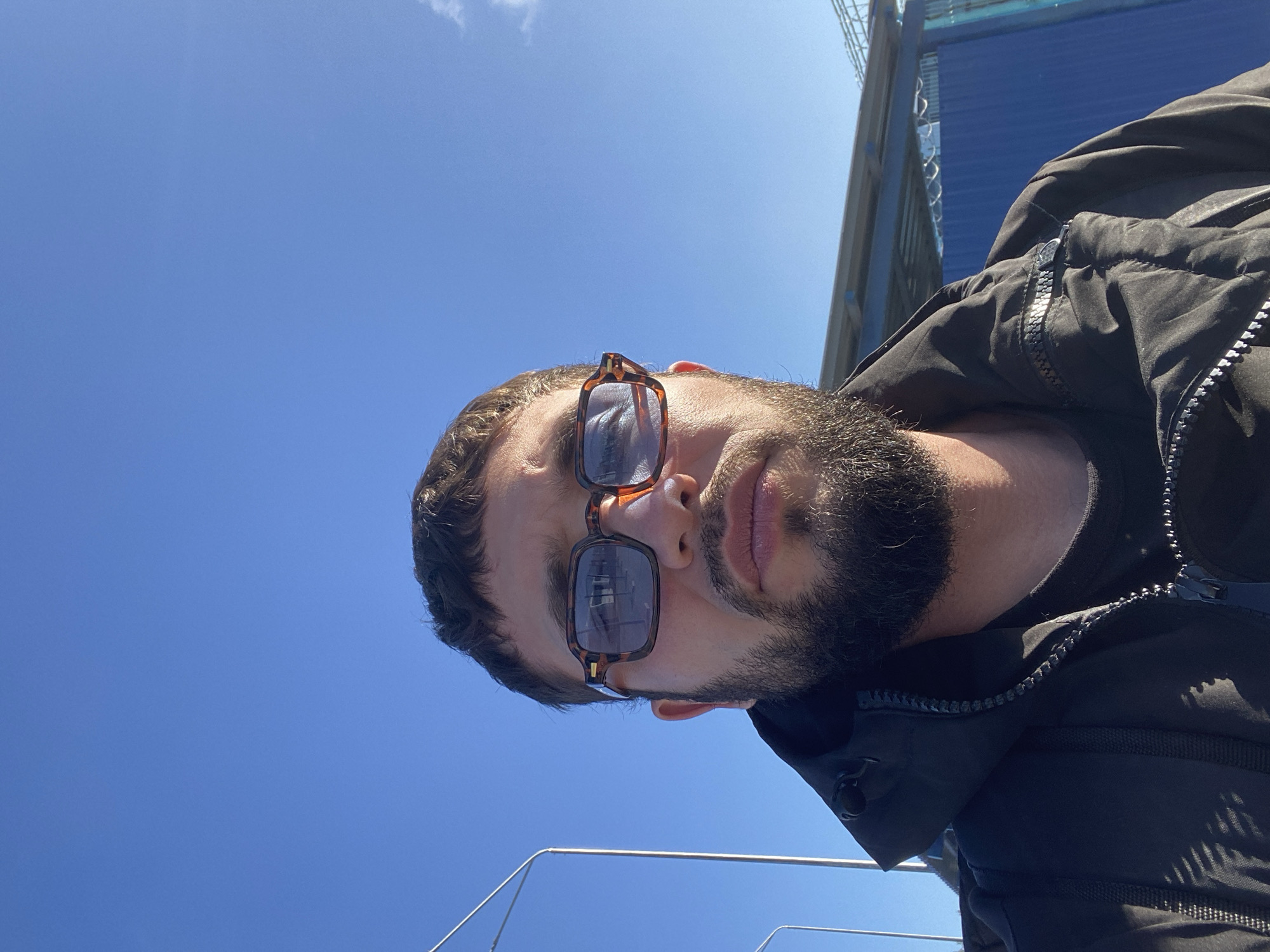 In a close-up, low-angle selfie, a man with dark hair and a beard, wearing tortoise-shell framed glasses and a black jacket, is pictured against a clear blue sky. The man's face is the primary focus, with the top portion of his head and upper body visible. In the background, the edge of a blue structure and what appears to be fencing or scaffolding are present. The overall lighting suggests it is daytime, with sunlight casting shadows. The location is identified as Koskolovo, Russia, though no specific landmarks indicative of that location are immediately apparent in the image itself.