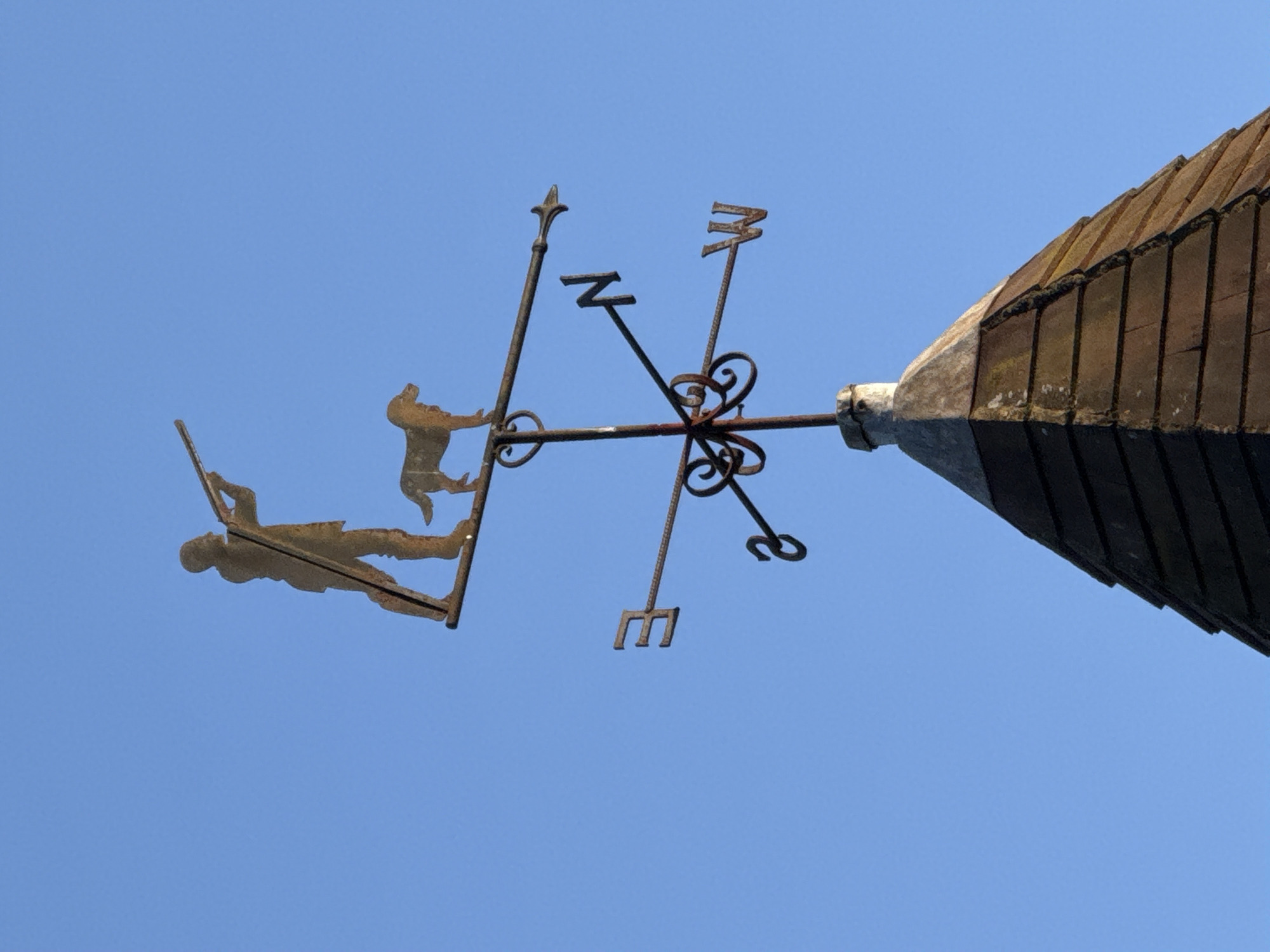 A low-angle, outdoor shot depicts a weathered metal weather vane, featuring directional letters (N, W, S, E) and silhouette figures, mounted on a building. The building, partially visible in the upper right, has a gray concrete or stone apex and a roof covered in dark brown, rectangular tiles. The weather vane's figures appear to depict individuals, one possibly reclining with an object in hand, the other standing. The sky is a clear, light blue, providing a uniform backdrop. The specific location in London, United Kingdom, is not ascertainable from the image alone.