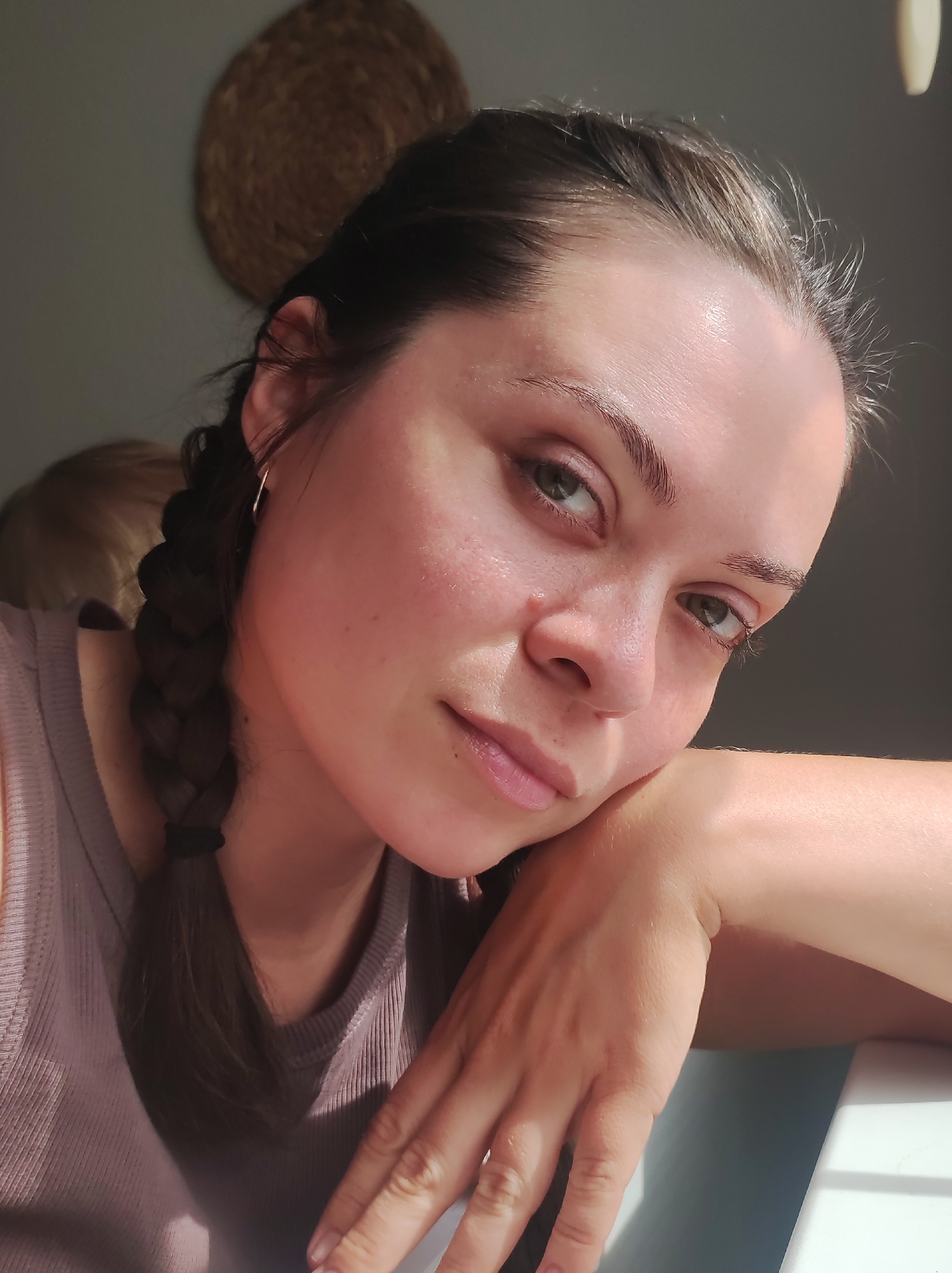 A young woman with dark hair in a braid rests her head on her arm, her gaze directed towards the viewer. She is wearing a light purple tank top and a subtle hoop earring. The lighting suggests it is daytime, with sunlight casting shadows across her face and illuminating her skin, revealing some texture. Her left arm is bent and resting on a surface that appears to be a white table. In the background, out of focus, is a wall with a round, woven decoration and the blurred form of another person. The overall scene is indoor, possibly a home, with a soft, natural light. There is no identifiable indication of the city of Chaikovsky, Russia within the image.