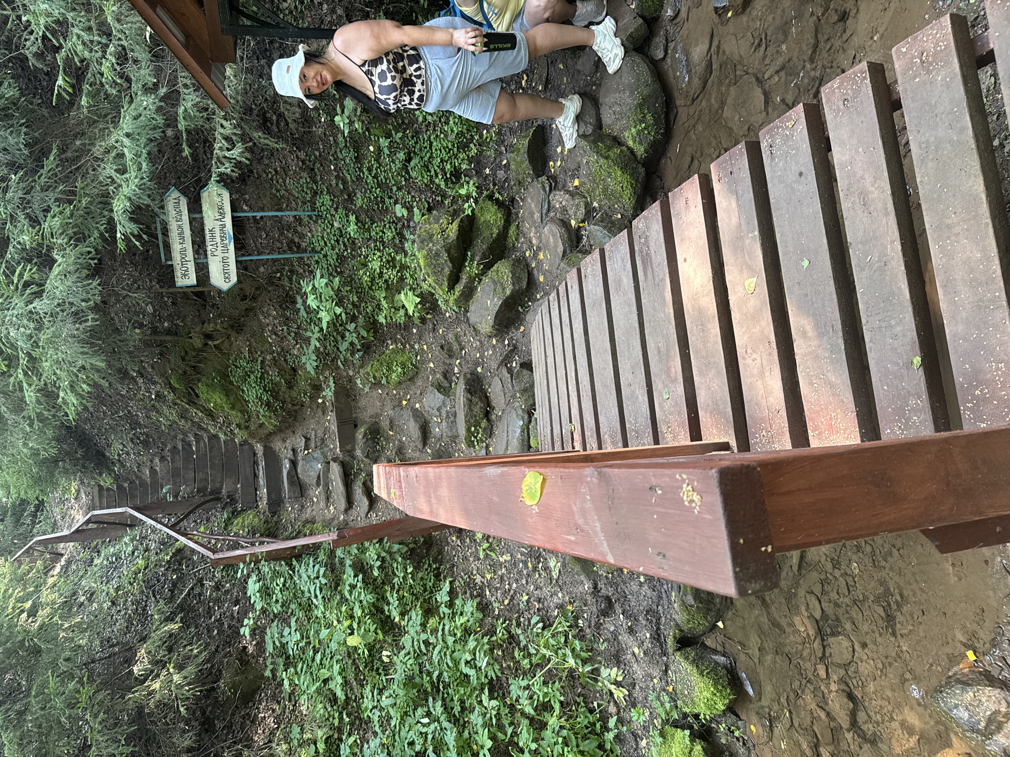 In Izhevsk, Russia, a wooden bridge spans a shallow, rocky stream in a wooded area. On the right, a woman wearing a leopard-print top, light blue shorts, and a white bucket hat stands on moss-covered rocks, holding a black water bottle. She is facing the camera and smiling. Behind her, a wooden structure with a roof is visible. Two directional signs are mounted on poles: one points left and reads "ЭКОТРОПА-КАНЬОН, ВОДОПАД" (Ecotrail-Canyon, Waterfall), and the other points slightly right and reads "РОДНИК СВЯТОГО ЦАРСТВЕННА АЛЕКСЕЯ" (Spring of Saint Tsarevich Alexei). To the left of the bridge, a steep wooden staircase with a metal railing ascends the forested hillside. The environment is lush with green foliage and trees.