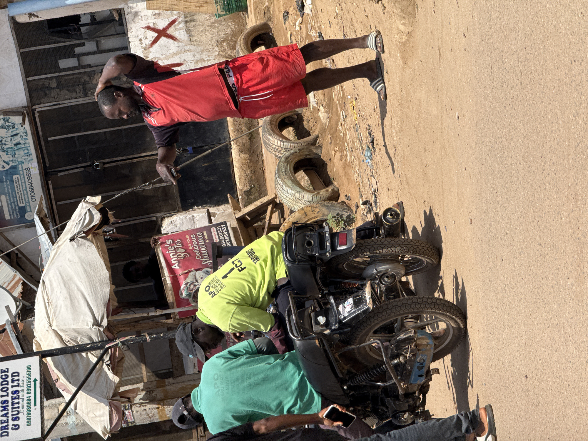 This image captures a street scene in Abuja, Nigeria, specifically near a roadside food stall and what appears to be a small commercial establishment. A man in a red and black polo shirt and red shorts, wearing striped sandals, stands with his hand on his head, looking to his left. He is standing on a dirt surface littered with debris, including several tires. In the background, a food stall with a white awning displays a sign for "Annie's Bites" advertising "Delicious Shawarma." Two men are seated on a motorcycle parked in front of the stall. One wears a bright green t-shirt with "APO RAINBOW" printed on the back, and the other wears a neon yellow t-shirt with various text and numbers, including "FCT." The motorcycle has a black seat and a visible license plate area. The overall environment suggests a bustling, informal commercial area within the city.