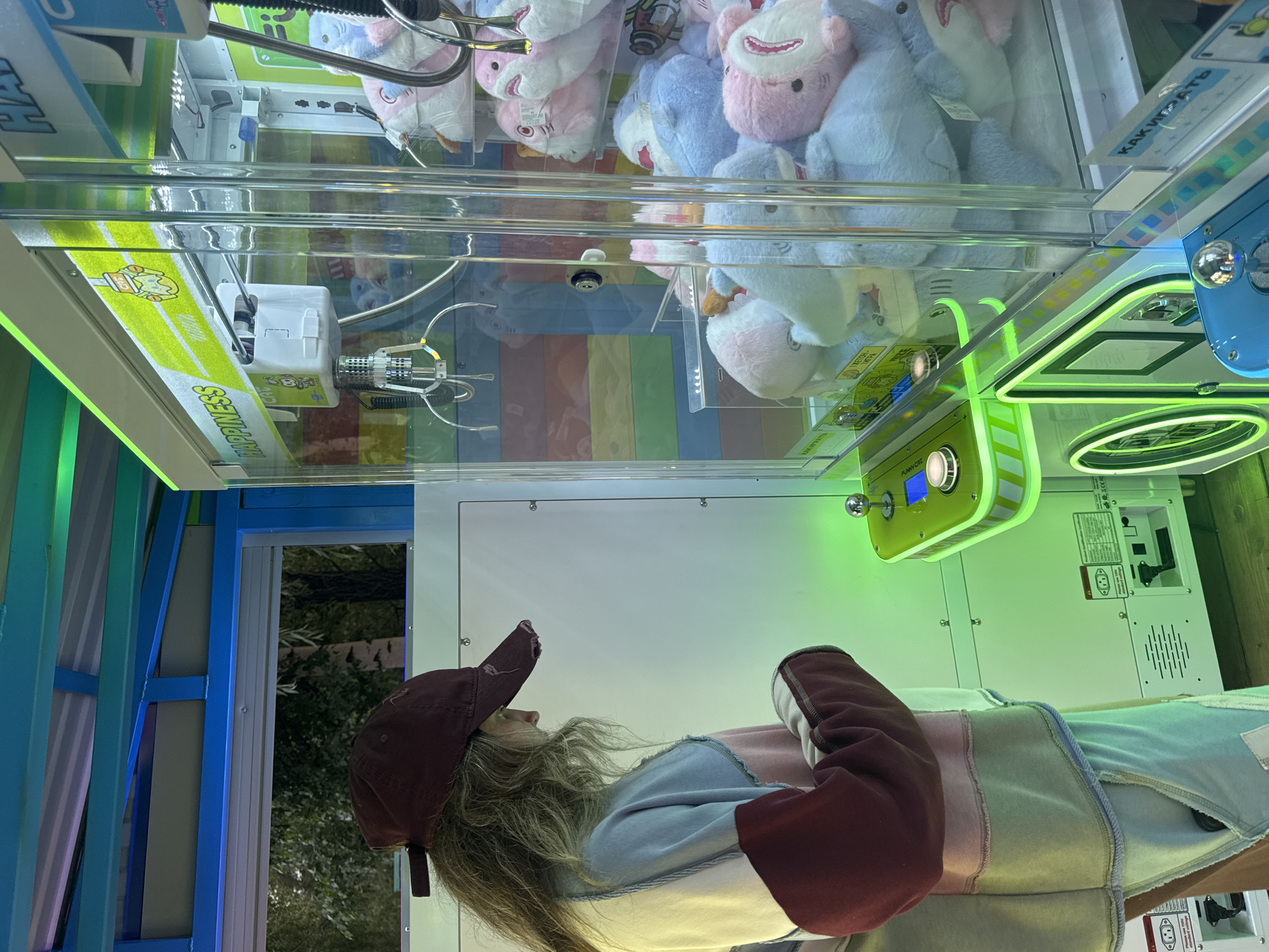 A young woman wearing a maroon baseball cap and a multi-colored sweatshirt stands in front of a claw machine arcade game. The machine is filled with plush toys, primarily pastel-colored sharks. The woman is looking down at the controls of the machine, which are lit with green LEDs. The background is a blurry outdoor scene with trees. The overall environment appears to be an amusement area, possibly an outdoor arcade. The image is taken from a slightly low angle, looking up at the woman and the machine. There are no other people visible in the frame. The scene is well-lit, with the green lights from the arcade game casting a glow.
