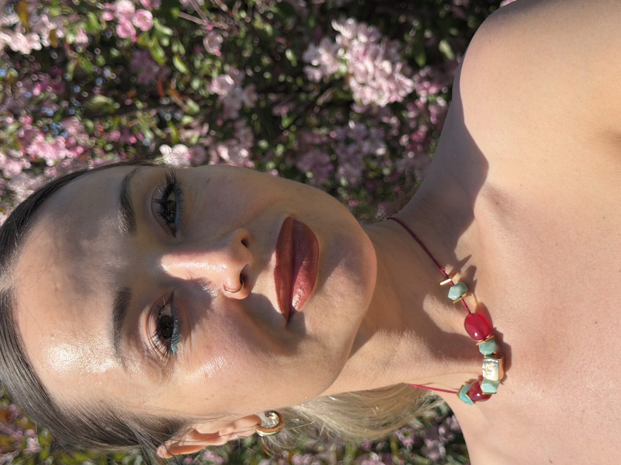 A close-up portrait features a woman against a softly blurred background filled with vibrant pink and purple blossoms, suggesting a springtime outdoor setting. Strong, direct sunlight illuminates her face and upper torso, creating distinct highlights and shadows, particularly on the right side of her face and shoulder. Her skin appears luminous and slightly dewy.
The woman has dark, defined eyebrows and wears winged eyeliner, which appears to be teal or dark blue. Her lips are adorned with a bold, reddish-brown lipstick. She sports a gold hoop nose ring in her left nostril and a small gold hoop earring in her left ear. Around her neck is a colorful necklace strung on a reddish cord, featuring alternating reddish-pink oval beads and light blue or turquoise irregular-shaped beads, with small gold-colored accents. Her light-colored hair is visible near her left ear.
The image captures her in a calm, composed pose, seemingly taken during a sunny day amidst blooming flora, typical of springtime in regions like Krasnoyarsk, Russia. There is no visible text in the image.