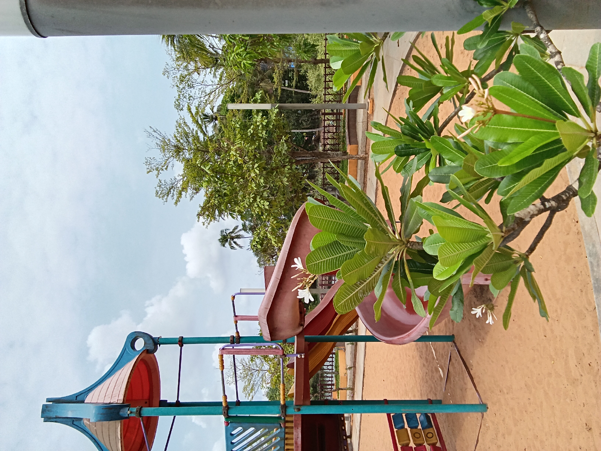 The image shows a playground scene in Chopdem, India. A brightly colored playground structure, including slides and climbing apparatus, is set on a sandy surface. Green trees and vegetation are visible in the background, partially obscured by a metal fence. In the foreground, branches with lush green leaves and white flowers frame the scene. The sky above is pale blue with scattered clouds. A grey pipe runs along the top edge of the frame.