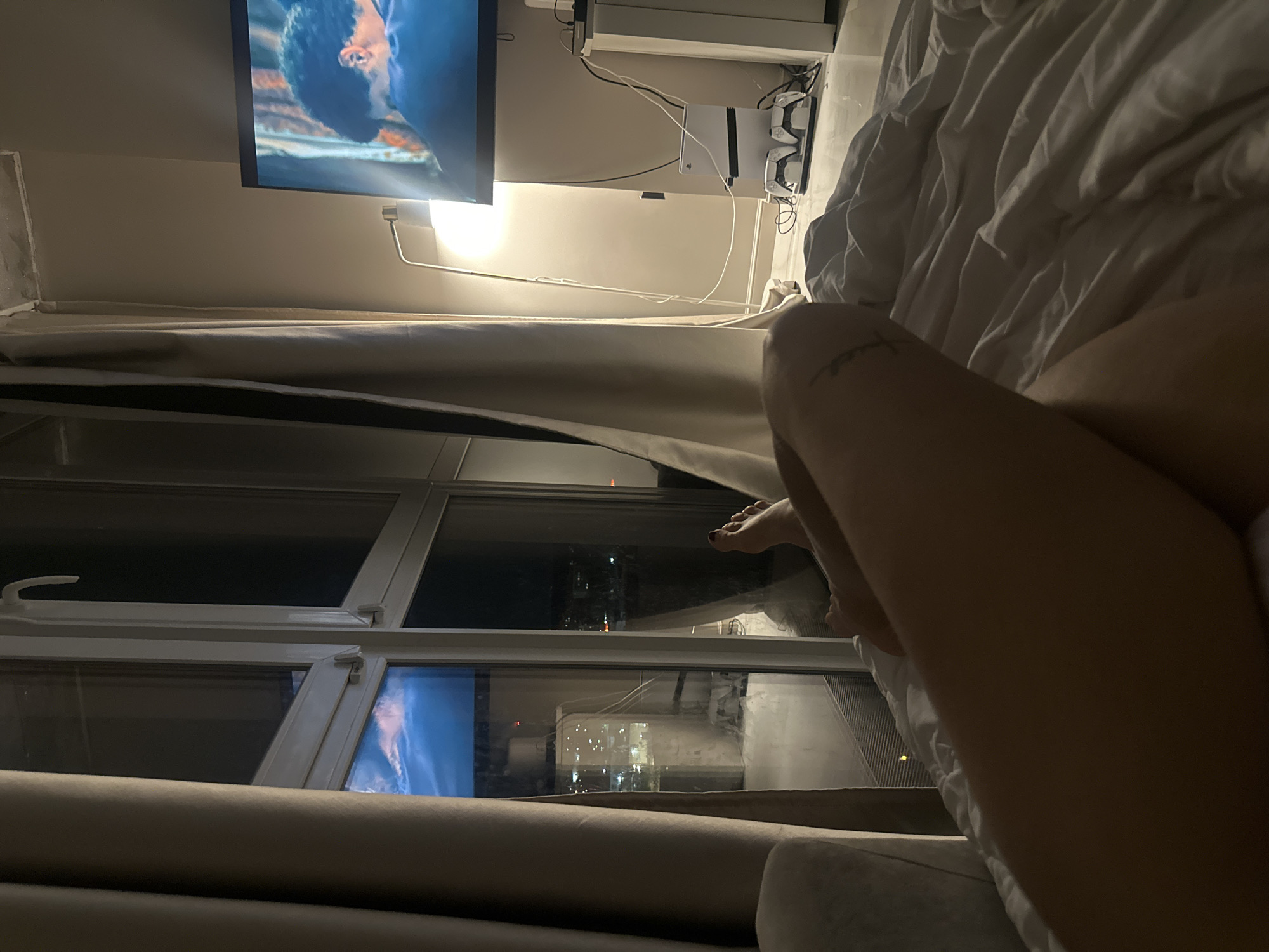 The image captures an indoor nighttime scene, likely from a bedroom or living room in Krasnogorsk, Russia. The perspective is first-person, showing a person's bare legs and feet in the foreground, suggesting they are relaxing in bed or on a couch. A small, indistinct tattoo is visible on the right leg, and one foot has dark nail polish.
In the midground, a large window reveals a dark night sky with reflections of city lights from buildings outside, confirming an urban setting. Partially drawn curtains frame the window. A television is mounted on the wall, displaying a close-up of what appears to be a person's face, possibly from a movie or TV show. Below the TV, a white PlayStation console (likely a PS5) is visible, accompanied by two white game controllers and a cluster of power cables. A flexible-arm lamp provides a warm glow to the area around the TV and console. The foreground also shows a rumpled white duvet or blanket, indicating a cozy, relaxed environment.
The overall lighting is dim, relying on the TV screen, the lamp, and the ambient glow from the city outside. The scene conveys a sense of quiet relaxation at night, potentially involving entertainment like watching TV or playing video games. No discernible text is visible in the image.