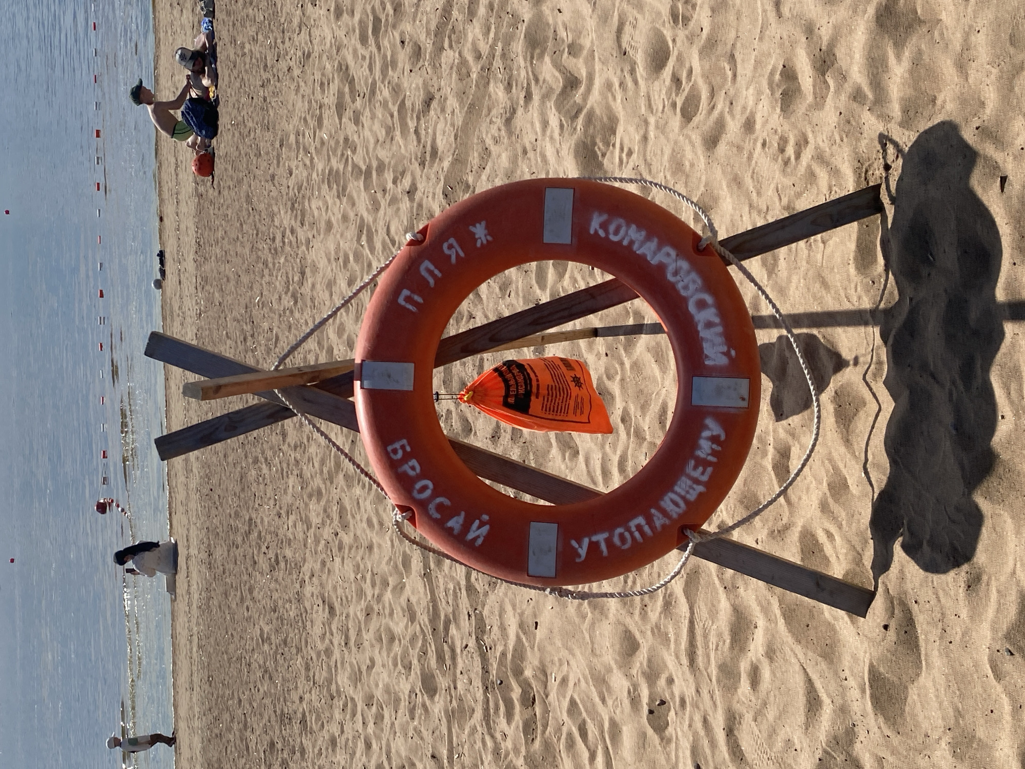 The image captures a sunny day at a sandy beach in Komarovo, Russia. In the foreground, an orange lifebuoy, resting on wooden supports, stands prominently on the sand. White Russian text is painted on the lifebuoy, reading "ПЛЯЖ КОМАРОВСКИЙ" (Komarovo Beach) at the top, and "БРОСАЙ УТОПАЮЩЕМУ" (Throw to the Drowning) at the bottom, indicating its safety purpose. A white rope is tied to the lifebuoy, and an orange, possibly inflatable, bag with some unreadable text in Russian hangs inside it.
The beach itself is wide and sandy, with distinct ripples and footprints. Several people are enjoying the day: two individuals are positioned on the sand near the water's edge, one sitting and another standing. Further along the beach, two children are playing in the sand, one wearing a hat. In the distance, other small figures can be seen at the water's edge or in the water. The water is calm, marked by a line of red and white buoys indicating a swimming area.
The scene suggests a leisurely summer day, with clear skies and bright sunlight. Shadows cast by the lifebuoy stand and people indicate the sun is at an angle, likely late morning or early afternoon. The overall impression is one of relaxation and recreation at a well-marked public beach.