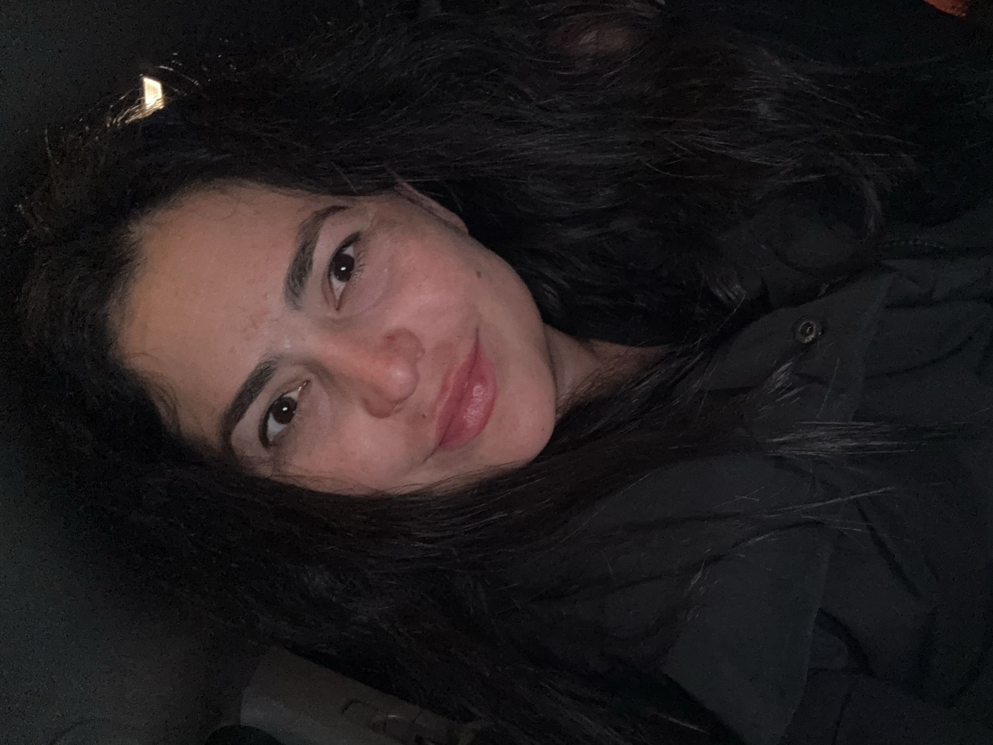 A young woman with long, dark hair is lying down, possibly in a vehicle, based on the interior and the angle of the shot. She is wearing a dark jacket. The lighting is somewhat dim. There is no information to suggest this image was taken in Kauchuk Stantsiya, Russia.