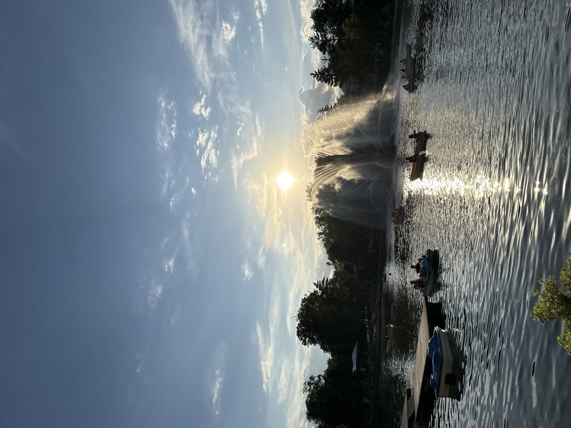 The image captures a vibrant outdoor scene on a sunny day, likely in a park in Moscow, Russia. The perspective is oriented sideways, but depicts a landscape view of a lake or large pond.
The dominant feature is a bright, low-hanging sun, positioned centrally on the left side of the frame (if rotated correctly). Its intense rays create a starburst effect and cast a strong glare on the water. A large, multi-stream fountain is prominently situated in the middle ground, directly in front of the sun, causing the water jets to glisten and appear backlit. Dark silhouettes of lush green trees line the far bank of the water body, framing the fountain and the sun.
The sky above is a clear, bright blue, adorned with scattered wispy cirrus clouds and some smaller cumulus clouds, suggesting fair weather.
On the right side of the image, the surface of the water is covered in ripples, reflecting the sun's light in shimmering streaks, creating a dazzling effect. Several recreational boats, including blue and white pedal boats and possibly yellow rowboats, are visible on the water. People are present in these boats, enjoying the leisure activity. Some boats are closer to a wooden dock or embankment in the foreground.
The overall atmosphere is one of peaceful recreation and natural beauty, characteristic of a pleasant late afternoon or early evening, given the angle and intensity of the sunlight. No discernable text is visible in the image.