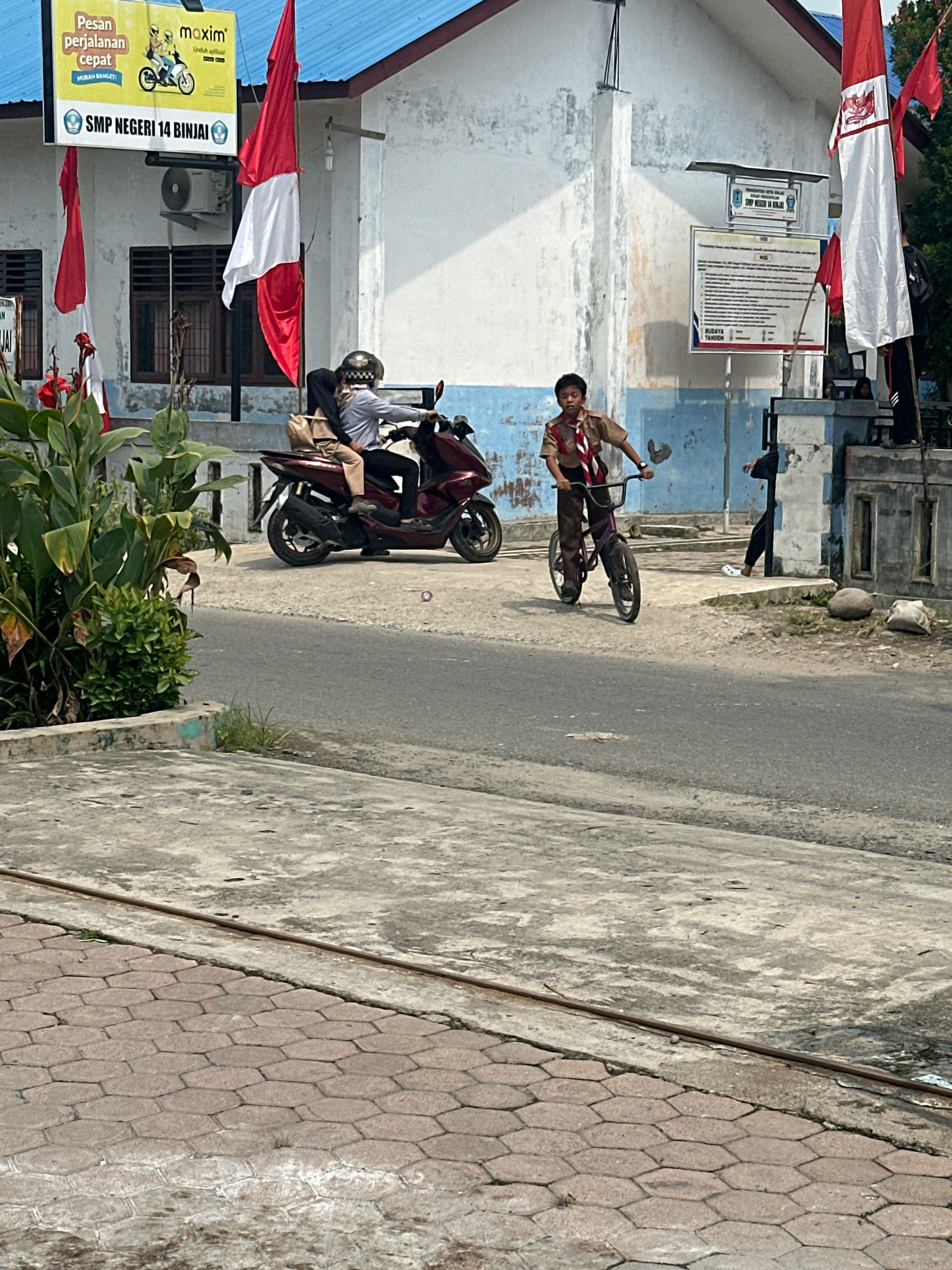 In front of SMP Negeri 14 Binjai, Indonesia, a young boy wearing a scout uniform rides a bicycle on a paved road. Behind him, two individuals are on a motorcycle, with the driver wearing a helmet. Several Indonesian flags are visible, including a large red and white flag and smaller red flags. The school building has a white facade with blue trim. The environment appears to be an urban street scene with concrete sidewalks and some vegetation.