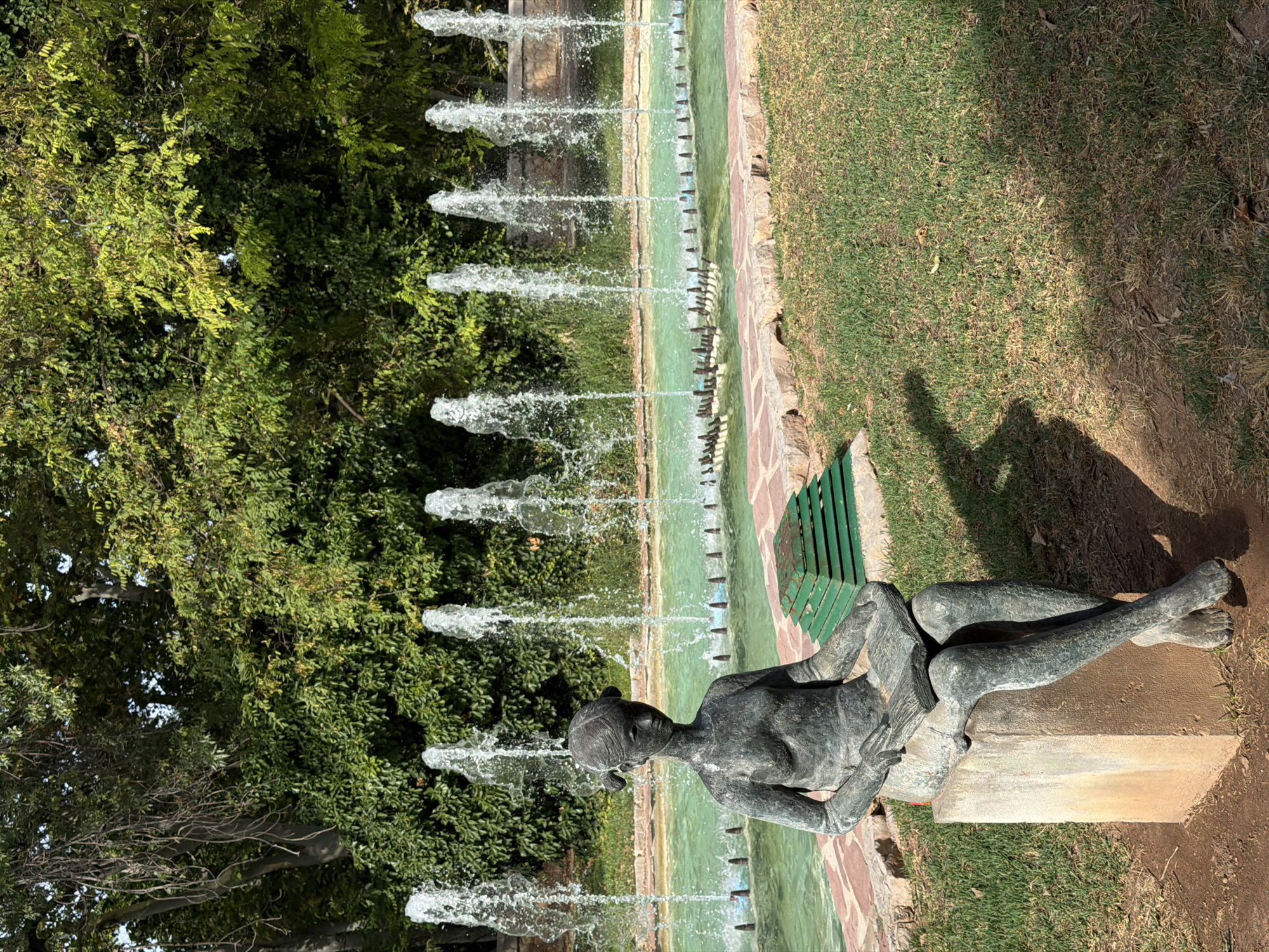 An outdoor scene features a bronze statue of a young girl sitting on a stone pedestal, holding an open book. She has her hair in pigtails and is looking down at the book. The statue is situated in a garden with a long, rectangular fountain in the background. The fountain has multiple jets of water spraying upwards into the air. Behind the fountain is a dense wall of green trees and foliage. The ground in front of the statue is covered in green grass, with some areas of dry grass and fallen leaves. The scene appears to be bathed in sunlight, casting shadows on the grass.