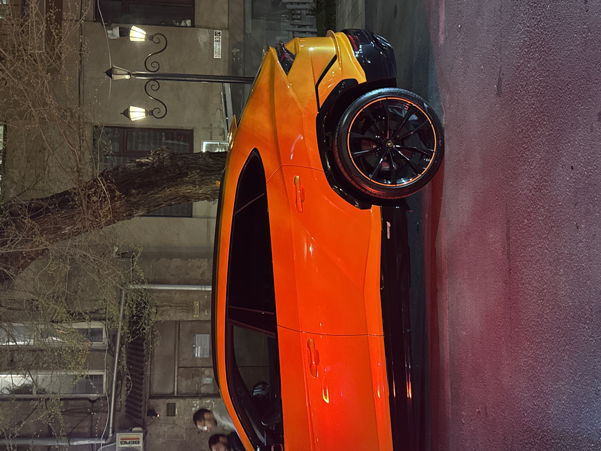 The image shows a parked orange and yellow Lamborghini Urus on a street in Yerevan, Armenia. The location is indicated by a building sign that reads "Nalbandyan 83/1" in Armenian. The car is parked on a paved road next to a building and a tree. There are two people near the front of the car. A streetlight illuminates the scene.
