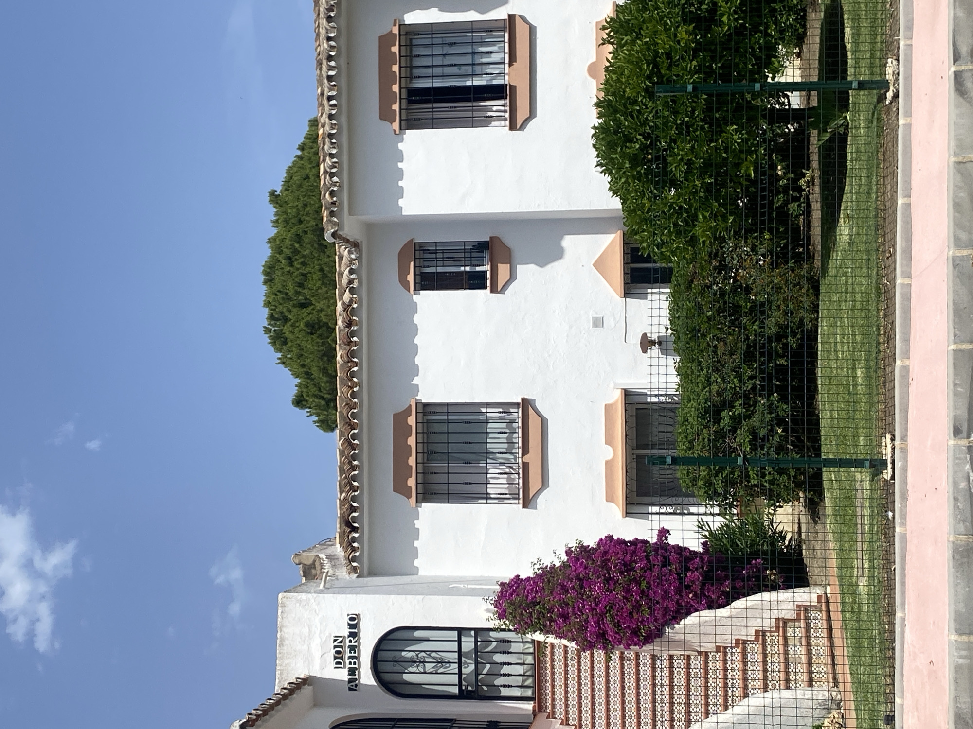 This is a medium shot, eye-level, outdoor photograph of a white building with a Spanish tile roof, located in Paraíso-barronal, Spain. The building features a prominent entrance on the left with a set of tiled stairs leading up to an arched doorway with a decorative metal gate. A vibrant purple bougainvillea bush is growing beside the stairs. Above the doorway, a sign reads "DON ALBERTO" in dark lettering. To the right of the entrance, the building has three windows, each with dark metal bars and decorative terracotta-colored awnings. Lush green bushes and trees are visible in front of the building, separated by a green metal fence. The sky is clear blue with a few scattered clouds.