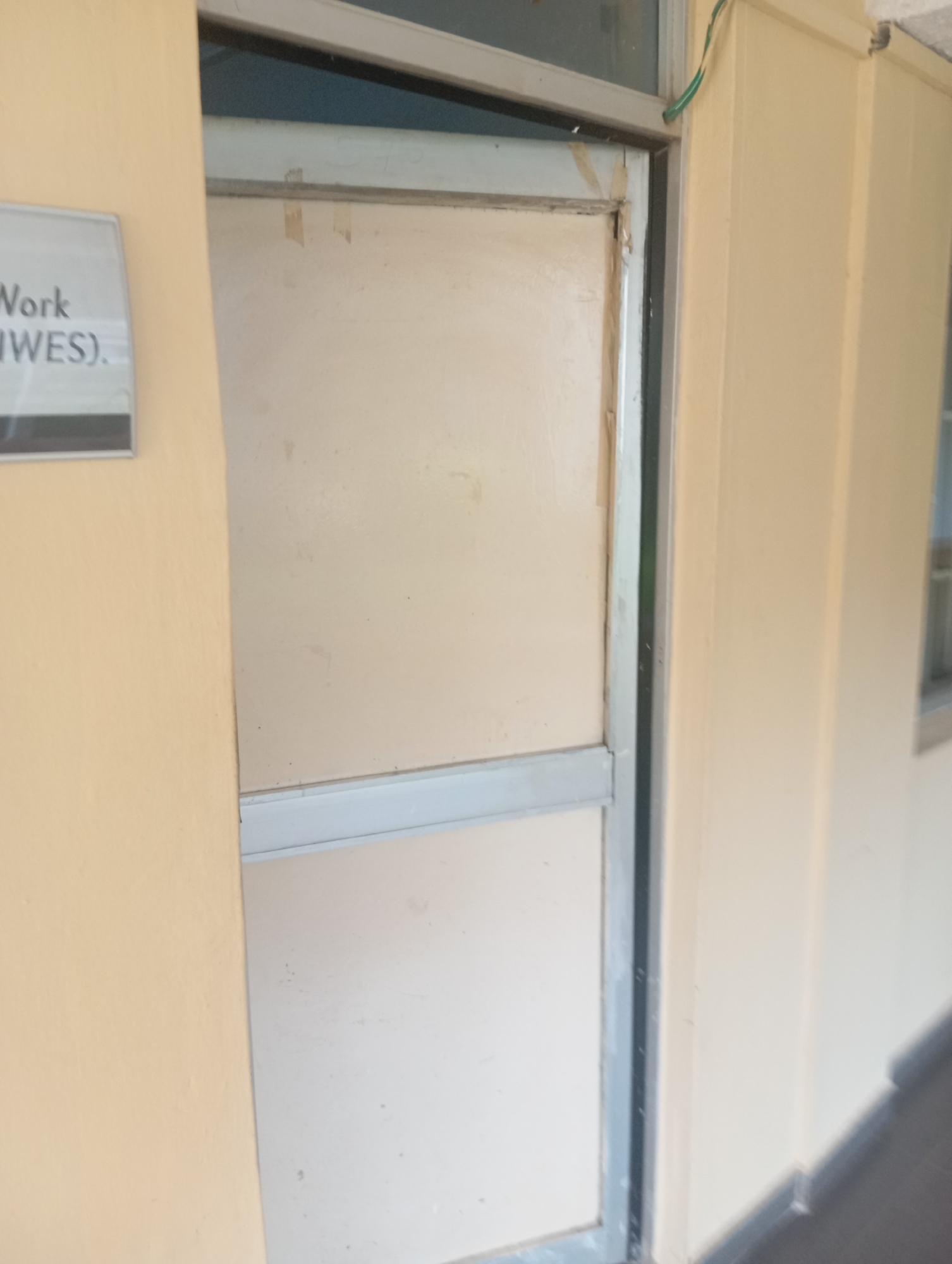The image shows a closed door with a light-colored surface, framed by a metallic border, situated in a hallway with light yellow walls. A sign on the left side of the door reads "Work (IWES)". A thin green wire is visible entering the top of the door frame from the right. The floor is not clearly visible, but a shadow suggests it is dark. The scene is likely an interior hallway of a building, potentially an office or educational institution, given the presence of the sign. The location is identified as Jos, Nigeria.