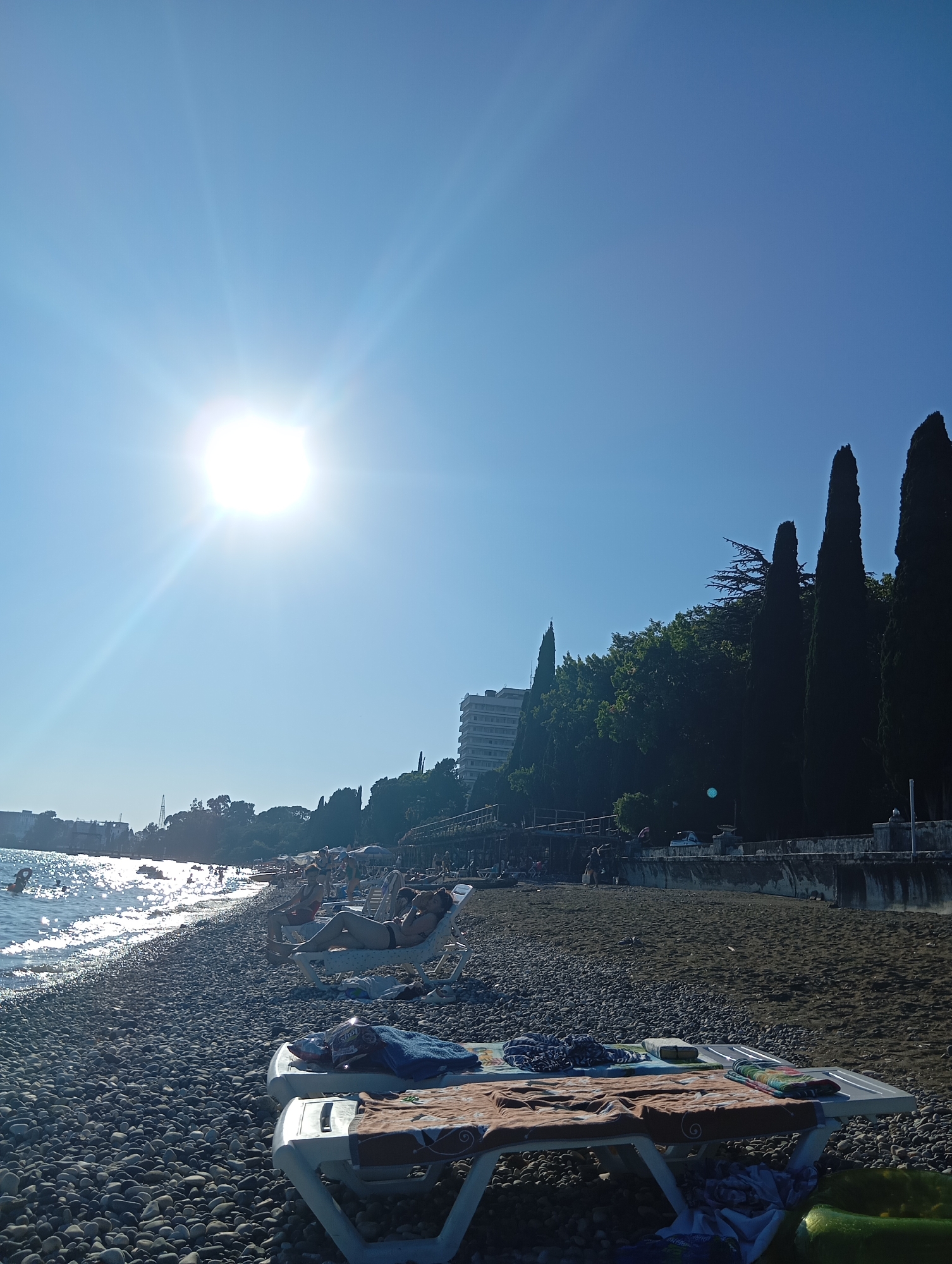 The image depicts a sunny day at a pebble beach in Sokhumi, Abkhazia. The sun is bright and high in the sky, casting a glare on the water. In the foreground, two empty sun loungers are covered with towels and clothing. A woman is visible reclining on a sun lounger further down the beach, wearing a bikini. The water is clear and reflects the sunlight, with several people swimming in the distance. Along the shoreline, there is a walkway with trees and buildings behind it. Tall, dark cypress trees are prominent along the embankment, silhouetted against the bright sky. The beach itself is a mix of dark sand and grey pebbles.