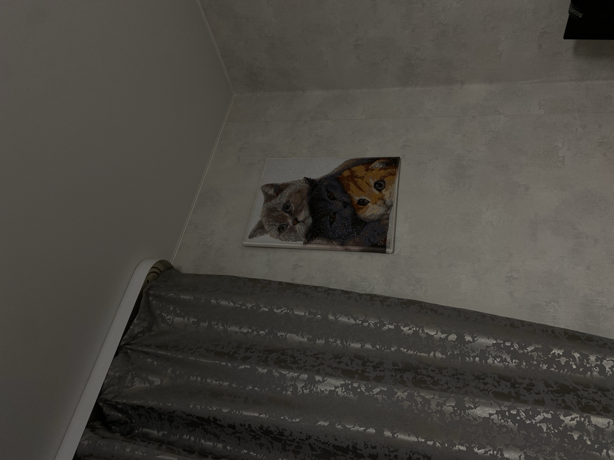 The image displays an indoor scene, likely a room in Chaikovsky, Russia, captured from a low angle looking upwards. The walls are light grey or beige with a subtle textured pattern. A corner where two walls meet is visible on the left side of the frame.
Centered on the upper wall is a framed artwork, appearing to be a diamond painting or a glittery print, which depicts two cat faces. One cat is light grey/brown, while the other is predominantly dark grey/black with some orange/brown fur visible on its face. Both cats have prominent eyes.
Below the artwork, filling the lower half of the image, are dark grey or black curtains. These curtains feature a textured, patterned design that reflects light, suggesting a metallic or shimmering effect. They are drawn or partially drawn, creating folds and ripples. A white, curved curtain rod or rail is visible along the left edge, supporting the drapes. In the upper right corner, a portion of a black electronic device is visible, with faint white text, possibly "SDES", on its side.
The lighting is dim and diffused, indicating either evening, night, or a poorly lit interior, contributing to a quiet and subdued atmosphere. No specific activity or event is taking place; the image is a static portrayal of the room's interior.