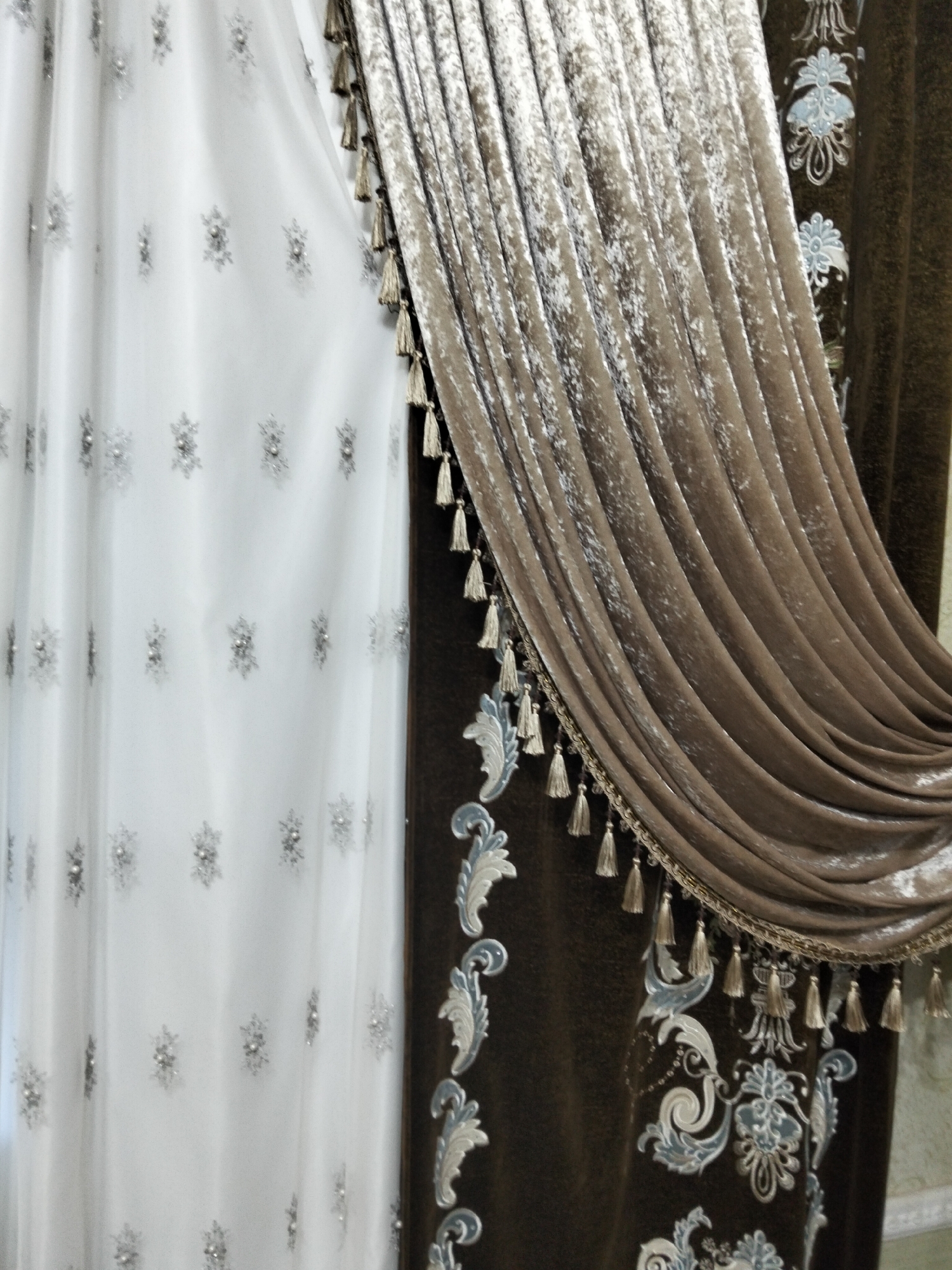 The image shows a close-up of two draped curtains. The left curtain is white and sheer, with a pattern of small, sparkling floral or snowflake-like embellishments. The right curtain is a rich, dark brown velvet with a textured sheen, gathered and draped to the right. This velvet curtain is adorned with a decorative trim along its bottom edge, featuring small tassels in a golden-tan color. The trim also includes a woven border. The velvet curtain itself is further embellished with an ornate embroidered pattern in shades of light blue and white, featuring scrollwork and floral motifs. The scene appears to be an interior setting, possibly a home or a commercial space, given the decorative nature of the curtains. There is no indication of the location being Kogon City, Uzbekistan; the image provides no geographical context.