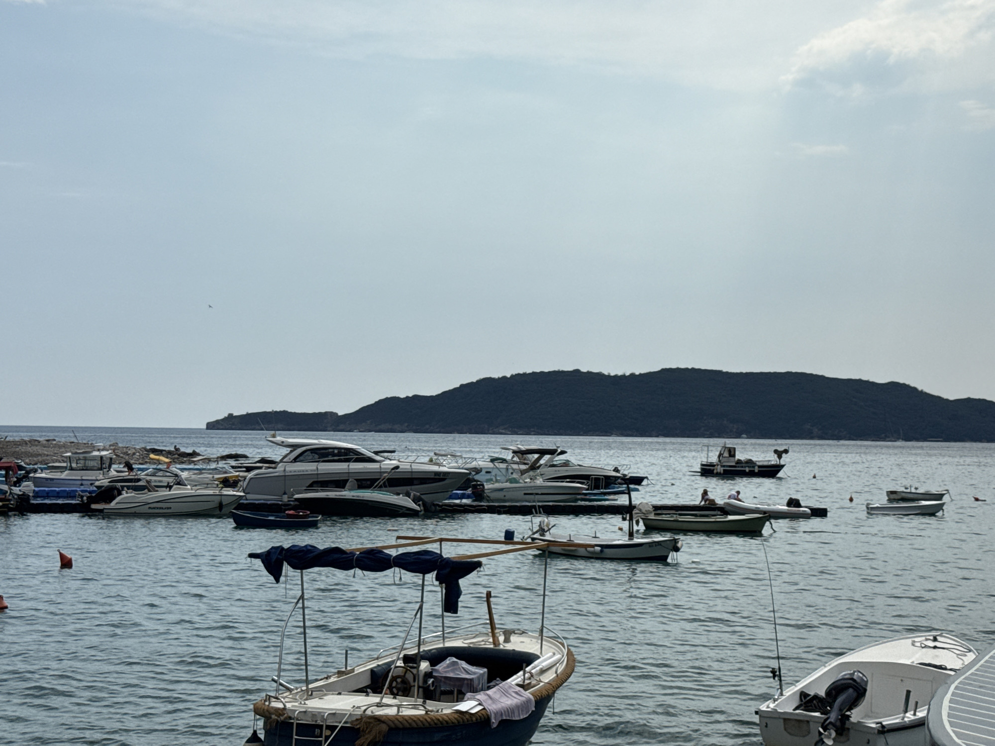 The image depicts a picturesque marina in Rafailovići, Montenegro. The scene is dominated by a variety of boats, ranging from sleek, modern yachts to smaller fishing vessels and dinghies, all bobbing gently on the calm, blue water. In the foreground, a small, weathered boat with a blue canopy is moored, its interior covered with a dark cloth.
Further out, several larger white motorboats and yachts are docked. Some have their canopies up, suggesting a sunny day. The background is framed by a lush, green, hilly island that slopes down to the sea, creating a dramatic backdrop. The sky is a soft, hazy blue with a few wispy clouds, indicating fair weather and likely midday or early afternoon. The water reflects the light, creating a serene and inviting atmosphere. There are no visible people actively engaged in an activity, suggesting a peaceful, leisurely scene. The overall impression is one of tranquility and maritime beauty.