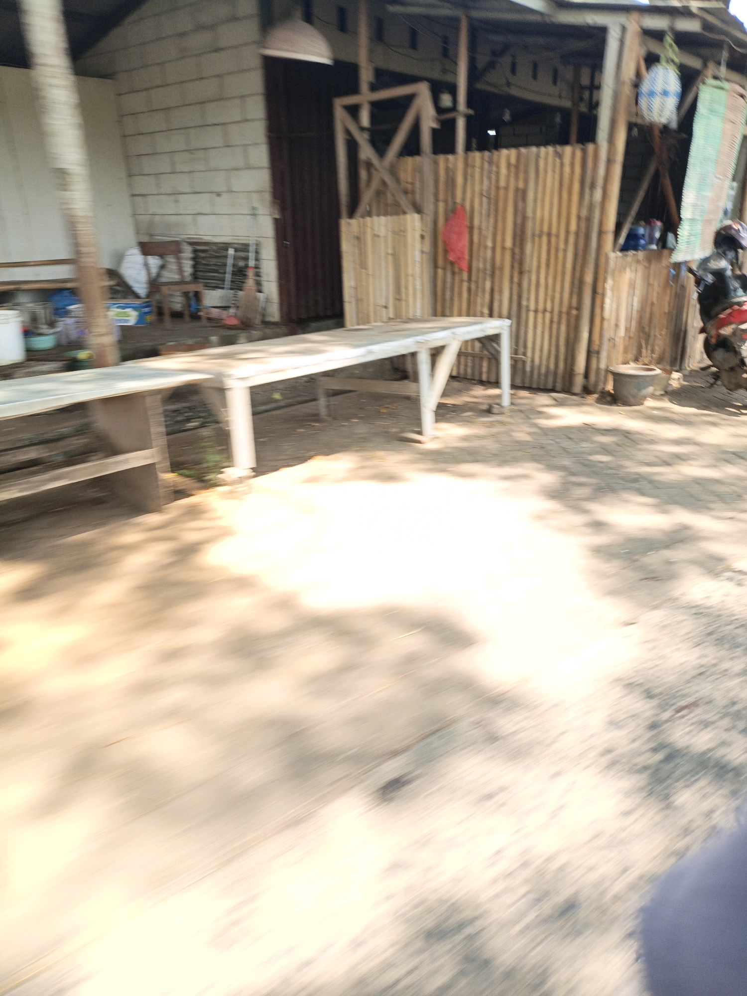 The image depicts an outdoor scene in Bandengan, Indonesia, featuring a makeshift structure constructed from bamboo poles and a long white table. The environment appears to be somewhat rural or semi-developed, with a dirt or gravel ground surface casting shadows from overhead foliage. In the background, a white brick building and a darker doorway suggest a dwelling or shop. To the right, a motorcycle is parked next to the bamboo structure, with a person wearing a helmet seated on it. The overall impression is one of a casual, perhaps temporary, gathering or vending spot.