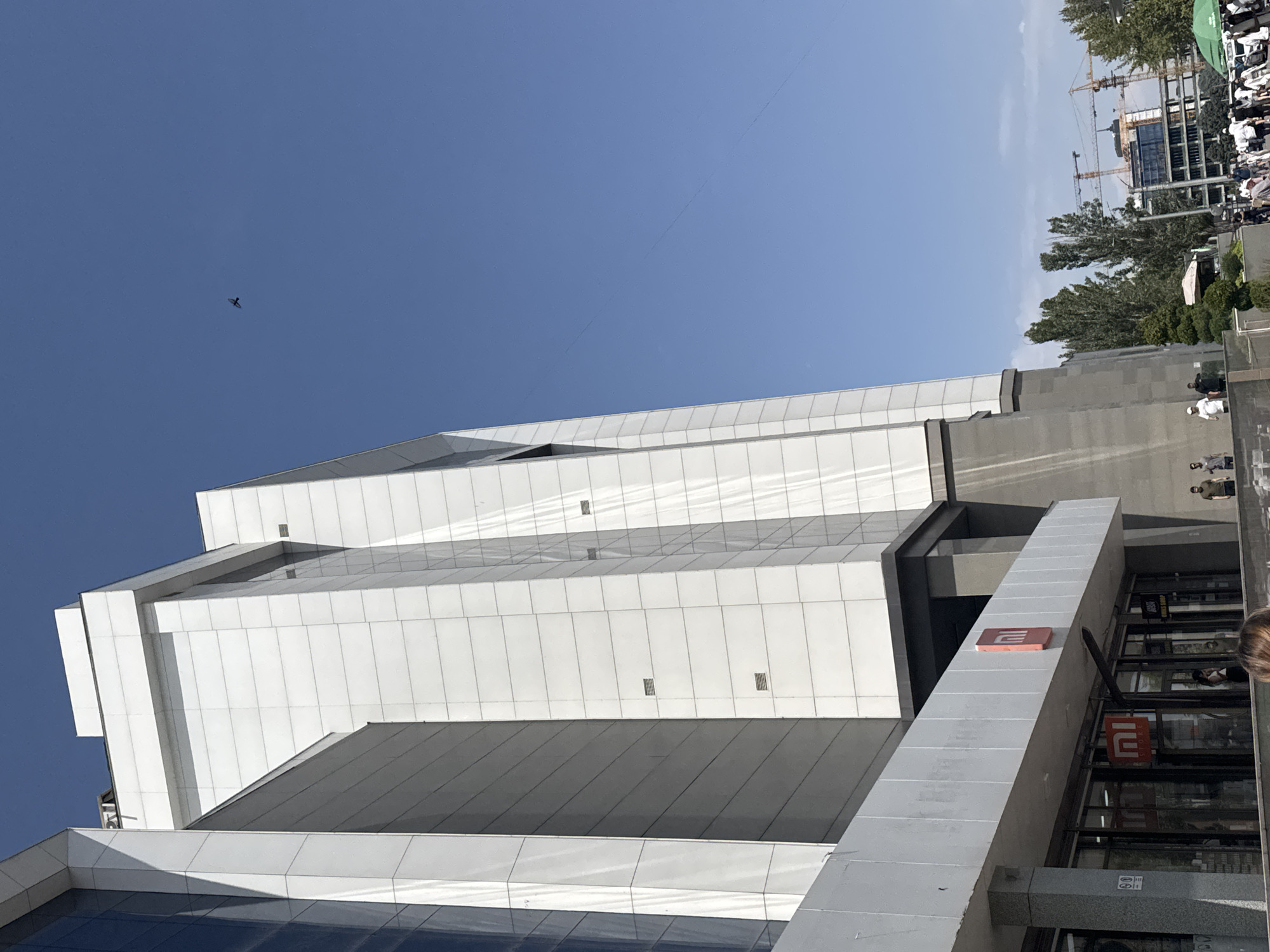 This is a ground-level, wide-angle, eye-level shot looking up at a modern white building with a glass facade on the left. The building has a sleek, geometric design. A prominent red "MI" logo is visible on a sign above an entrance. The sky is clear blue with a small dark object, possibly a bird, in flight. In the background, trees and construction cranes indicate an urban environment. Several people are visible in the foreground and middle ground, some walking and others standing. The overall scene suggests a sunny day in a developed urban area. The building appears to be a commercial establishment, possibly a store. The presence of the "MI" logo strongly suggests this is a Xiaomi store. Based on available information, the Xiaomi store at Bishkek City Mall in Bishkek, Kyrgyzstan, features a similar architectural style and logo.