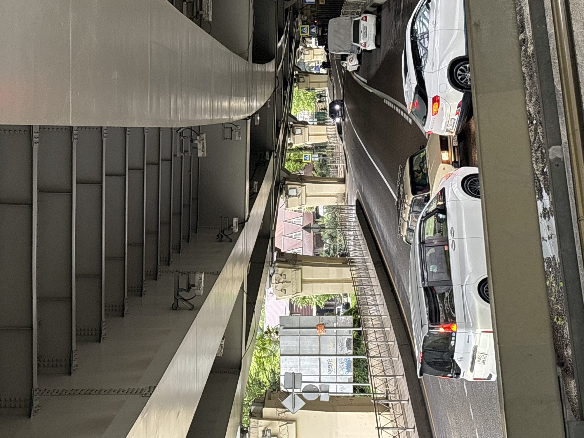A vertical, high-angle shot depicts a street scene beneath a bridge, likely in Sochi, Russia. Traffic moves on the asphalt road; the vehicles include white cars and a van. The bridge's metal support structures dominate the left side of the frame, showing the engineering of its construction. Signs and buildings are partially visible between the bridge supports, adding depth to the scene. The lighting is natural, suggesting daytime.
