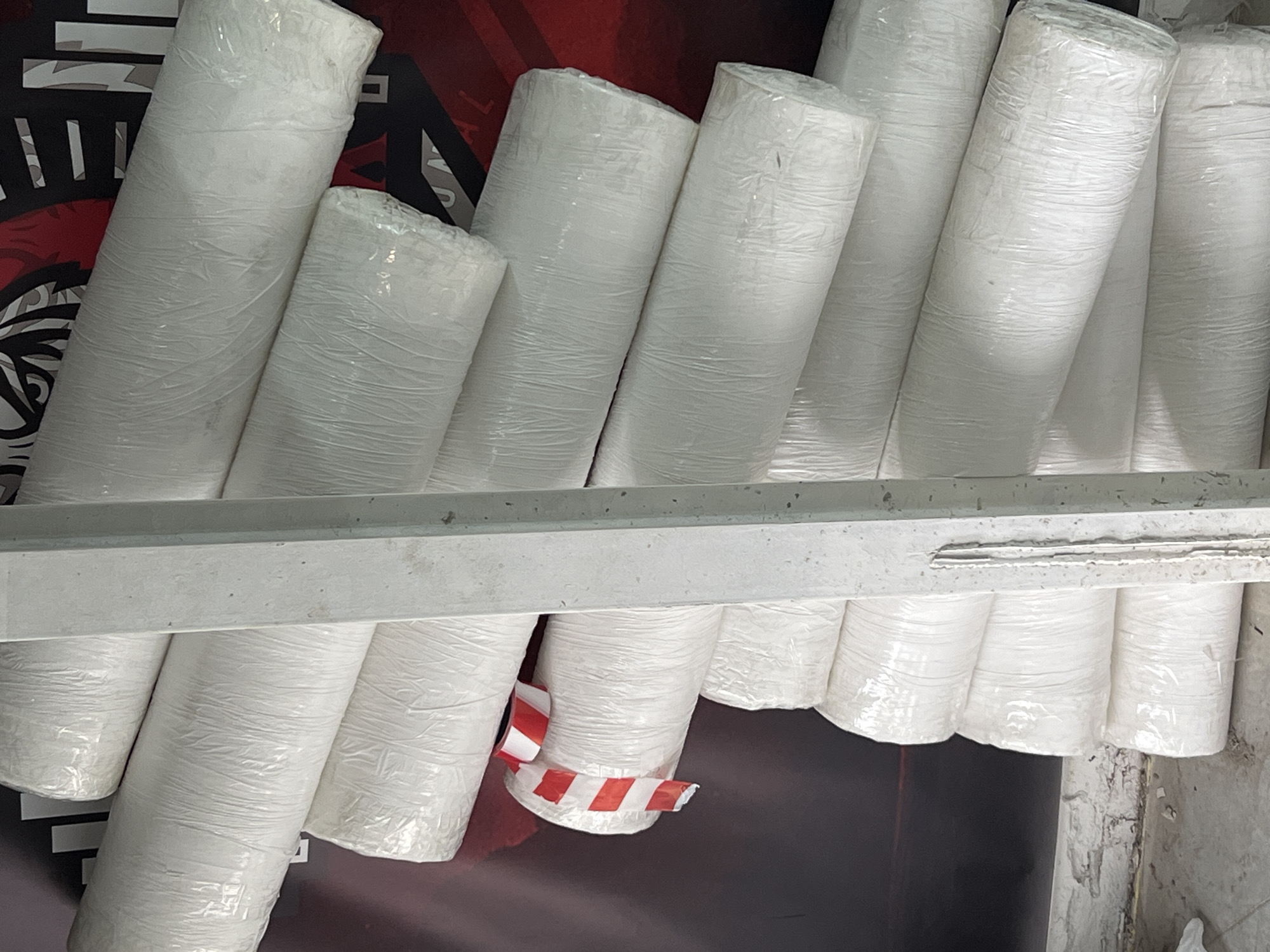 The image captures a scene featuring numerous large rolls of white material, each encased in clear plastic wrapping. These cylindrical rolls are stacked vertically and leaned against one another, filling a significant portion of the frame.
A grey metal shelf or beam is visible horizontally across the mid-section of the image, appearing to separate two tiers of these stored rolls. In the lower section, one of the rolls is distinctly marked with a piece of red and white striped warning or caution tape wrapped around its base.
The background behind the rolls consists of a dark red surface with abstract black patterns. Partial white text is visible on this background, with the letters "AL" and other indistinct characters being discernible, possibly forming part of a larger word such as "MATERIAL" or a brand name.
The setting suggests an indoor environment, likely a warehouse, storage facility, or a shop stocking bulk items, given the industrial appearance of the metal shelving and the packaging of the goods. There are no people present, and no specific activity is taking place beyond the storage or display of the materials. The lighting appears even, typical of an indoor space, and there are no cues to indicate the time of day or outdoor weather. The overall impression is one of organized inventory.