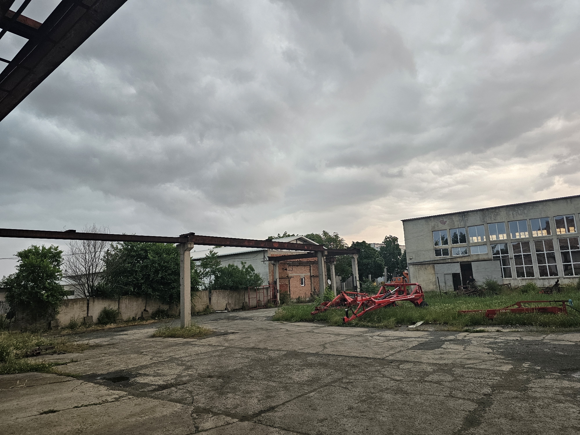This image depicts an overcast outdoor scene of a disused industrial or agricultural yard in Karnobat, Bulgaria. The foreground is dominated by a cracked concrete ground, showing wear and tear, with numerous small puddles and patches of overgrown grass and weeds pushing through the cracks.
In the mid-ground, a prominent, rusty, dark brown metal gantry or frame structure spans horizontally across the scene, supported by large, grey concrete pillars. Behind this structure, lush green trees and bushes grow, partially obscuring other buildings. On the far left, parts of a light-colored building are visible behind the foliage. Towards the center-back, a brick building with a small rectangular window can be seen.
On the right side of the image, a large, multi-story, grey building stands. It features numerous windows, some reflecting the cloudy sky, while others appear grimy, broken, or boarded up. The building's facade is discolored and shows signs of significant neglect. In front of this building, nestled among tall green grass and weeds, lies a piece of red agricultural machinery, possibly a hay rake or similar farm implement, appearing abandoned. Another smaller red metal object is visible further to its right.
The sky is heavily overcast with dense, grey clouds, indicating a dull day, possibly after rain given the puddles on the ground. A faint patch of lighter sky is visible towards the upper right, hinting at obscured sunlight. The overall atmosphere conveys a sense of abandonment, disuse, and industrial decay. There are no people visible, and no discernible activity is taking place. No legible text can be identified on any of the objects or buildings.