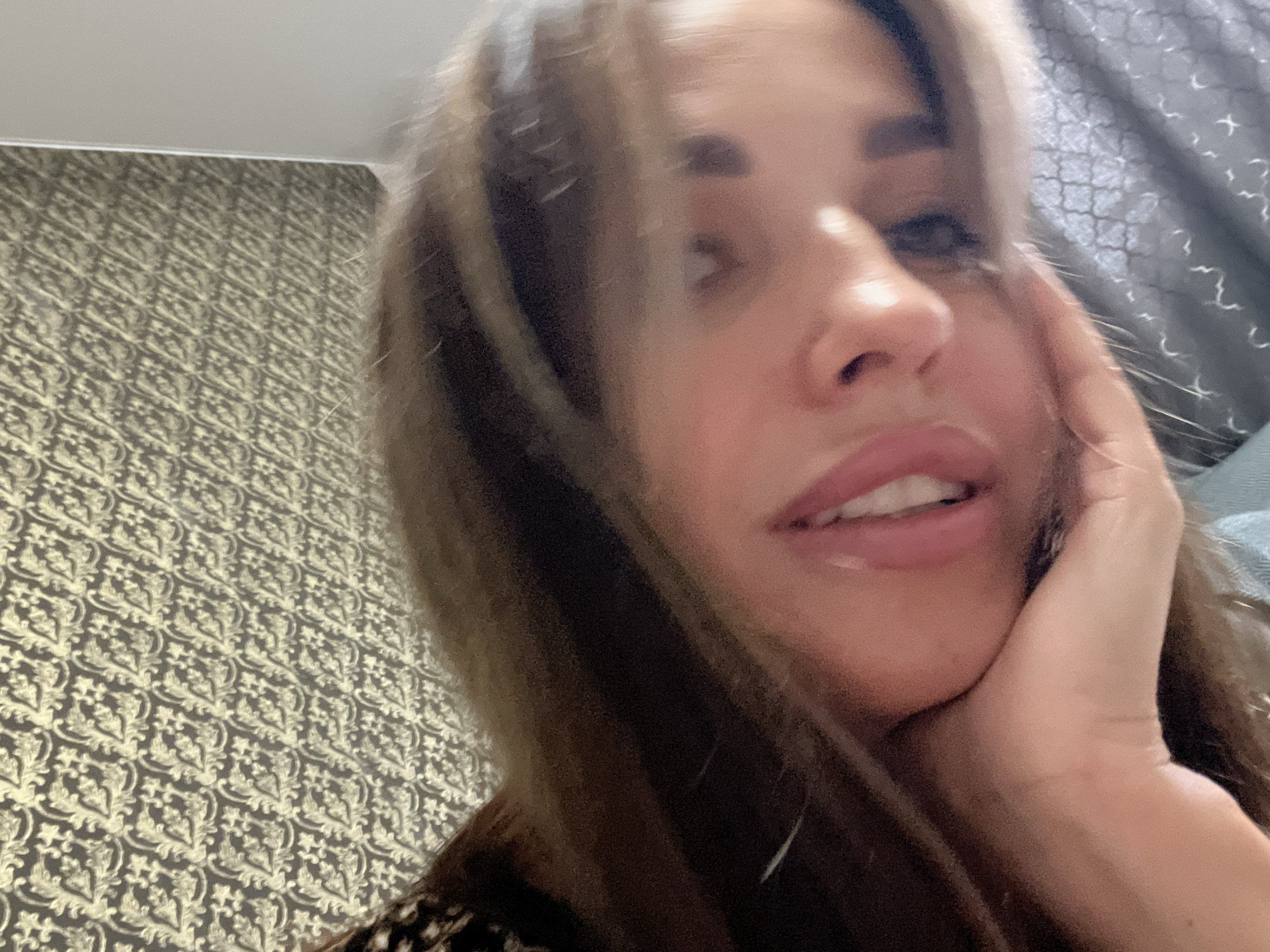 A close-up, slightly blurred, high-angle selfie shows a person with long, light brown hair resting their head on their hand. The background consists of a wall with a textured, damask-like pattern in gold and dark brown, and a portion of a bed with a gray, patterned duvet. The person's face is turned towards the camera, with their mouth slightly open, revealing teeth. Their eyes are not clearly visible due to the blur and angle. The lighting suggests an indoor setting.