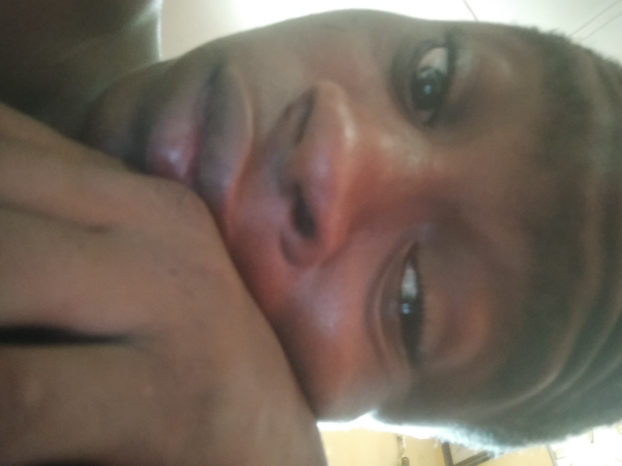 A close-up self-portrait of a Black male is presented, focusing on his face from a low angle. His left hand is positioned near his mouth, partially obscuring it. The background is indistinct, but the setting appears to be indoors, potentially in a residential space. The photograph was reportedly taken in Shangisha, Nigeria.