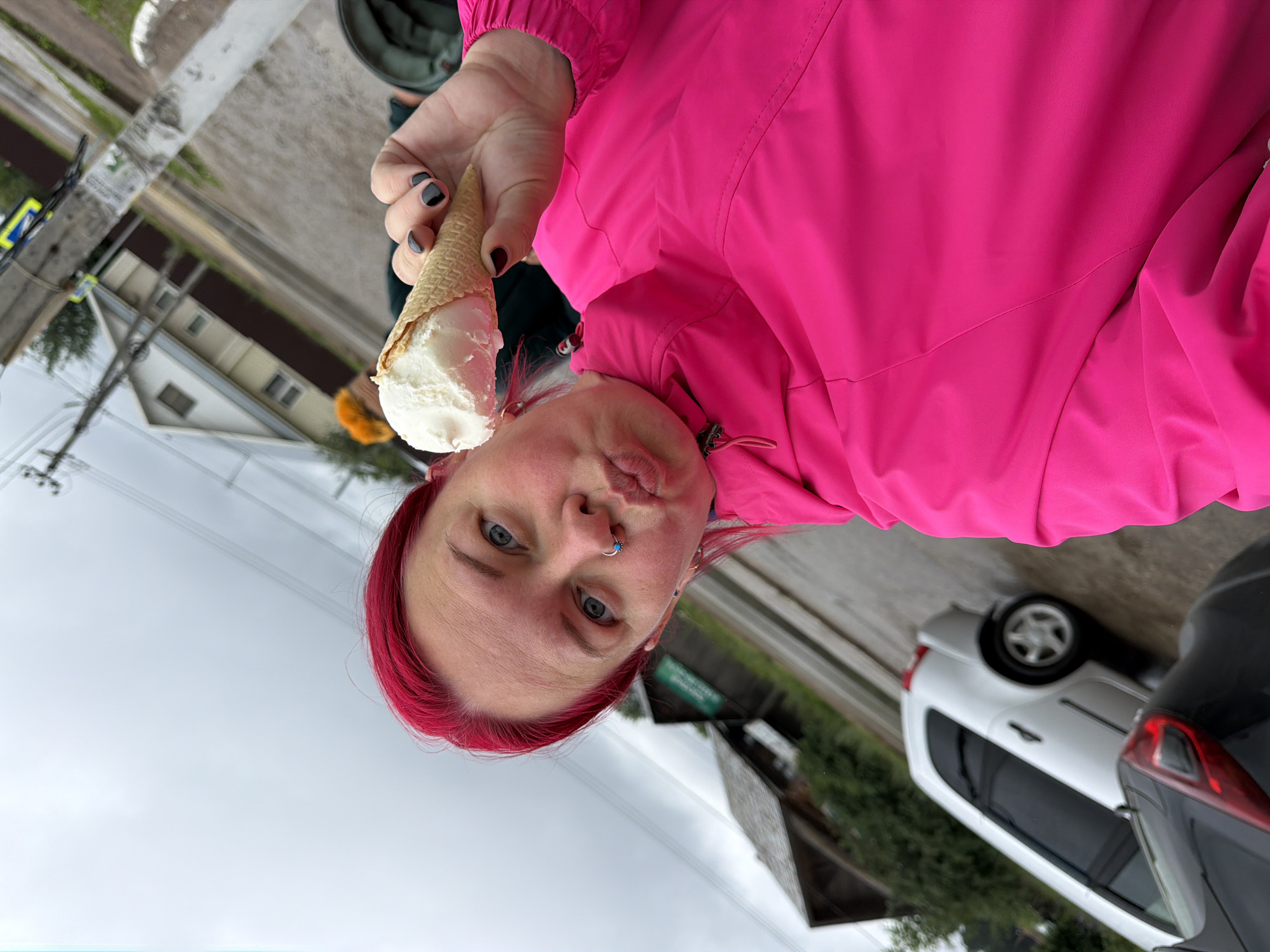 A woman with bright pink hair is taking a selfie while holding an ice cream cone. She is wearing a vibrant pink jacket. Her facial expression suggests she is about to kiss the camera. Behind her, a white car is visible, and in the background, there are houses and trees, indicating a residential or semi-rural setting. The sky is overcast. The context suggests this is an outdoor scene in Chaikovsky, Russia.