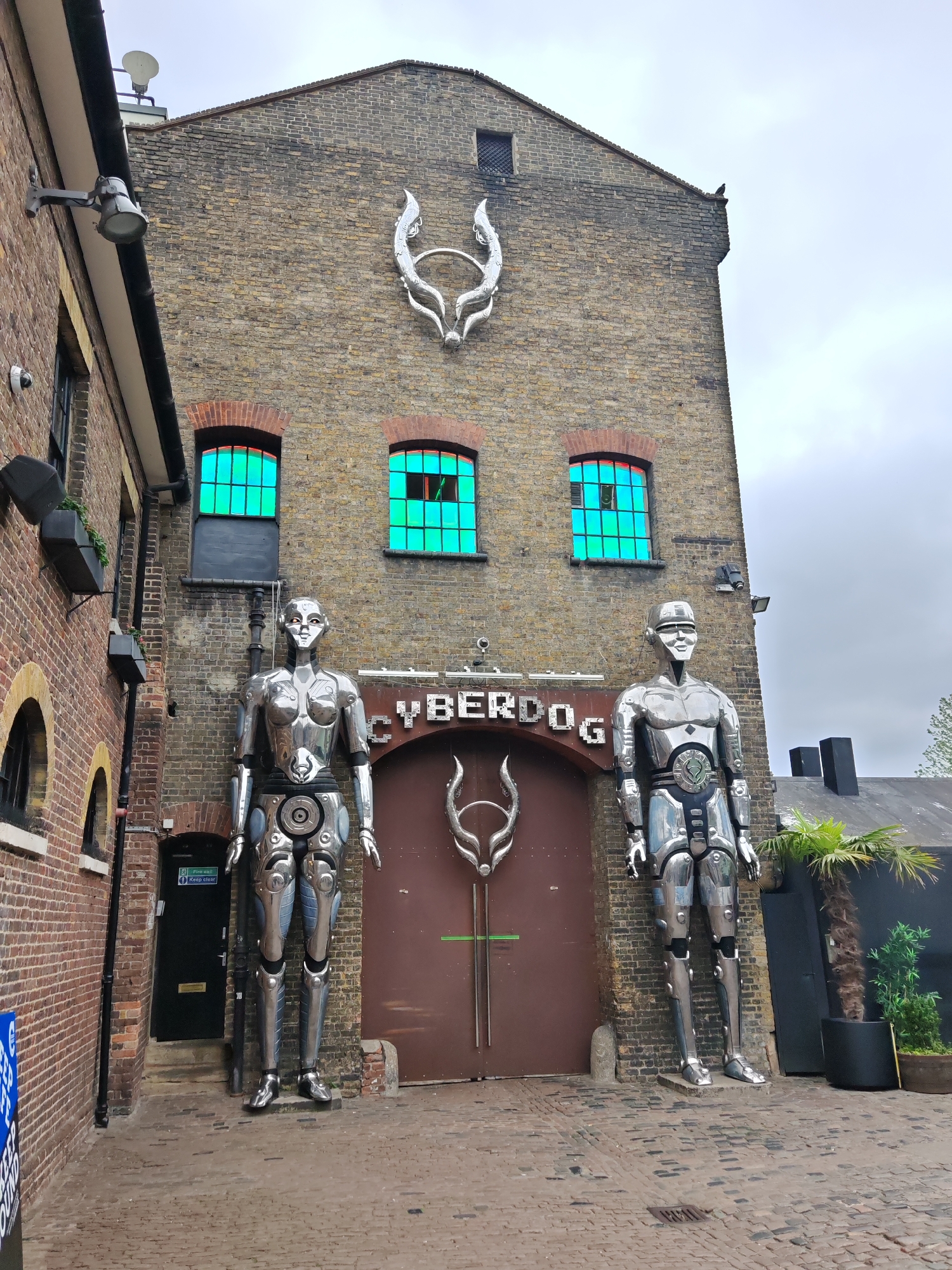 The image shows the exterior of Cyberdog, a shop known for its futuristic and rave-themed clothing, in Camden Town, London, United Kingdom. The building is a multi-story brick structure. Above the shop entrance is a sign reading "CYBERDOG," flanked by two large silver humanoid robot statues, one male and one female. The entrance itself is a large, dark-colored door with a silver logo resembling antlers. Above the entrance on the building's façade are three windows with vibrant green frames. A similar silver logo is mounted higher on the building. To the left of the Cyberdog entrance is a brick building with arched windows and a black door with signage. To the right is a black structure with palm trees in pots. The sky is overcast. The foreground is a cobblestone walkway.