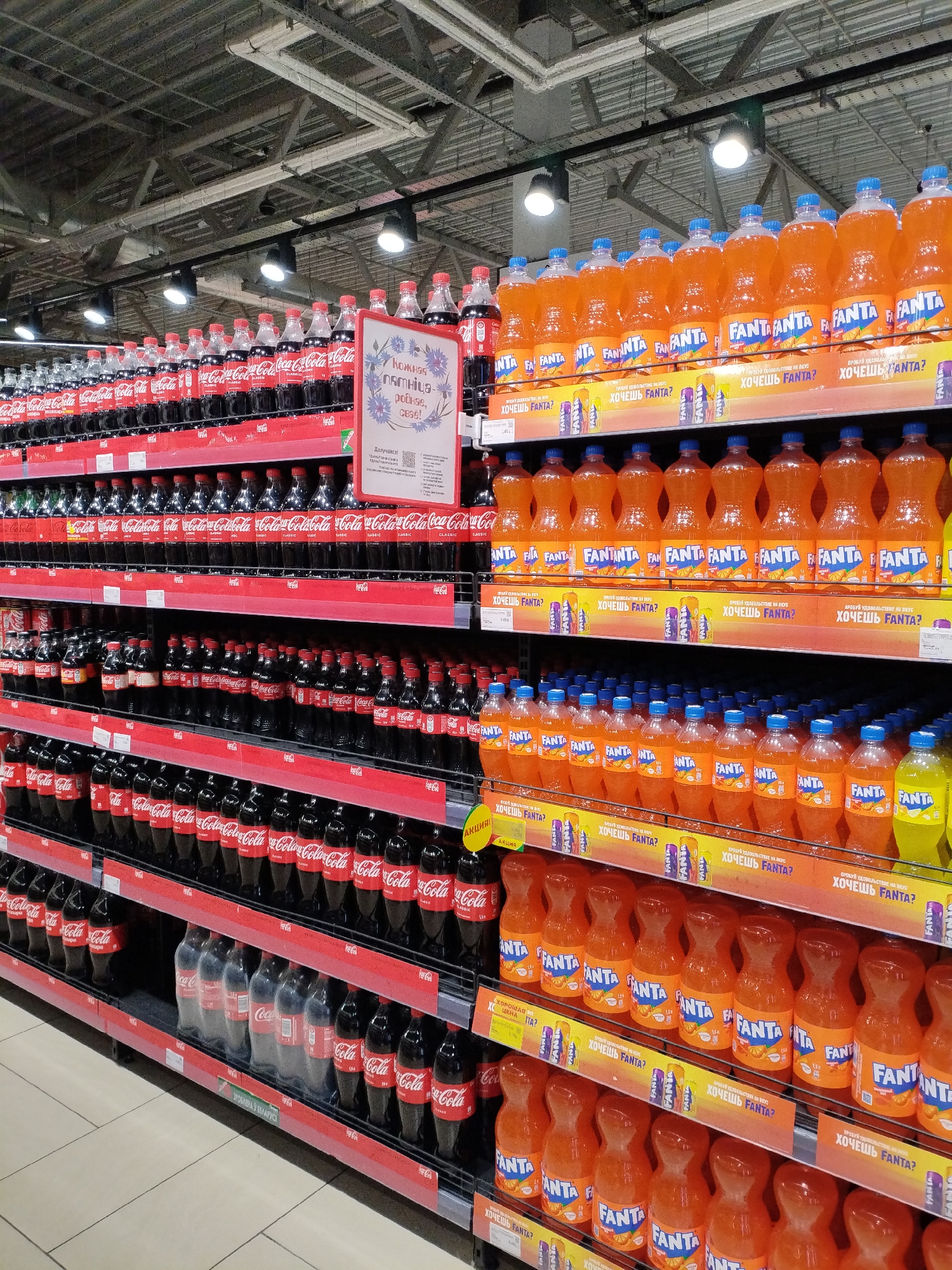 The image shows shelves stocked with various soft drinks in a supermarket in Mahilyow, Belarus. The predominant products are Coca-Cola and Fanta. The shelves are organized by brand, with rows of bottles neatly arranged. Some shelves display multiple tiers of the same product. Promotional signage, primarily in Russian and featuring the Fanta brand, is visible on some shelves, indicating sales or special offers. The lighting appears to be from overhead fluorescent fixtures. The overall scene is a typical retail environment for beverages.