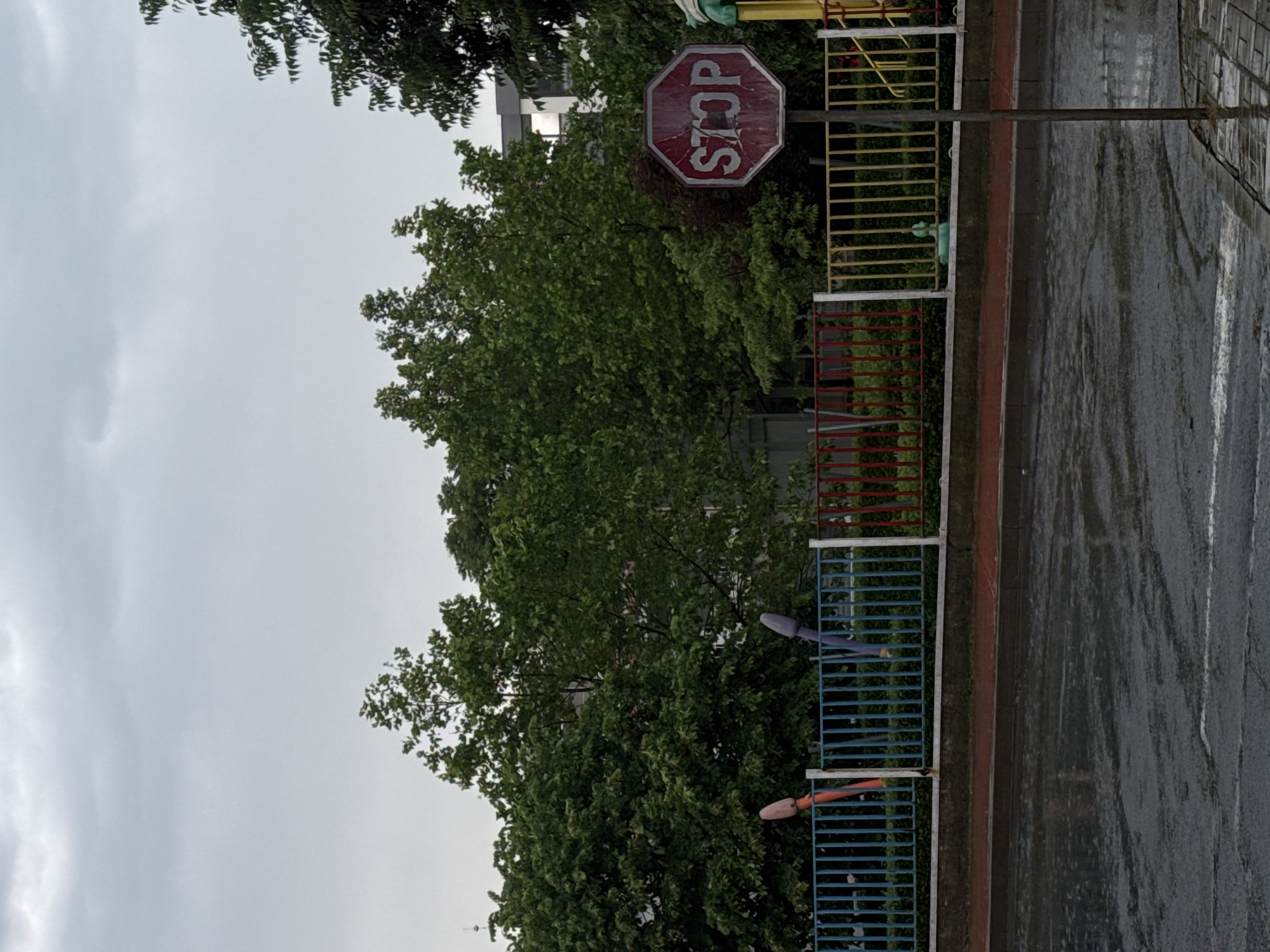 This image, captured in Tsarevo, Bulgaria, presents an outdoor scene on an overcast day, likely after recent rain, indicated by the wet asphalt. The perspective is slightly angled, looking towards a roadside or pathway.
In the mid-ground, a weathered red octagonal stop sign is prominently displayed, featuring the word "STOP" in white capital letters. The sign is mounted on a metallic pole, extending vertically from the ground. Behind the stop sign and fence, lush green trees dominate the background, their dense foliage suggesting a park or forested area. A building is faintly visible through the trees behind the sign.
In the foreground, running parallel to what appears to be a wet road or paved area, is a vibrant metal fence. This fence is divided into distinct sections, painted bright yellow, red, and blue. On the blue section, two decorative, elongated shapes, possibly wind chimes or stylized fish, are attached – one purple and one orange. The fence separates the road from the dense greenery.
The sky is a uniform, light grey, completely covered with clouds, indicating an overcast day. There are no people or animals visible, and no specific activity is taking place, rendering the scene quiet and still.