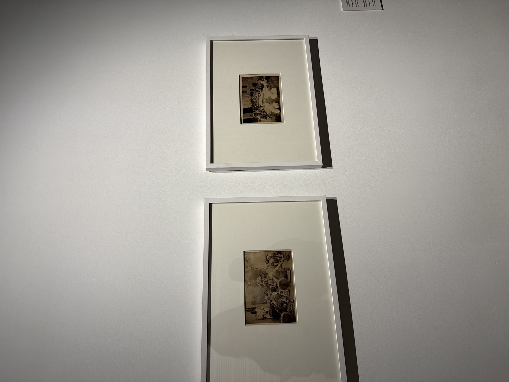 Two framed black and white photographs hang on a white wall. Each photograph is small and centered within a larger white mat and framed in a thin white frame. The top photograph appears to depict people or figures in a historical setting. The bottom photograph also seems to show a group of people in a vintage scene, possibly involving some sort of equipment. The photographs are spaced vertically, suggesting they are part of a gallery or museum exhibition. The location is unknown and not discernible from the image.