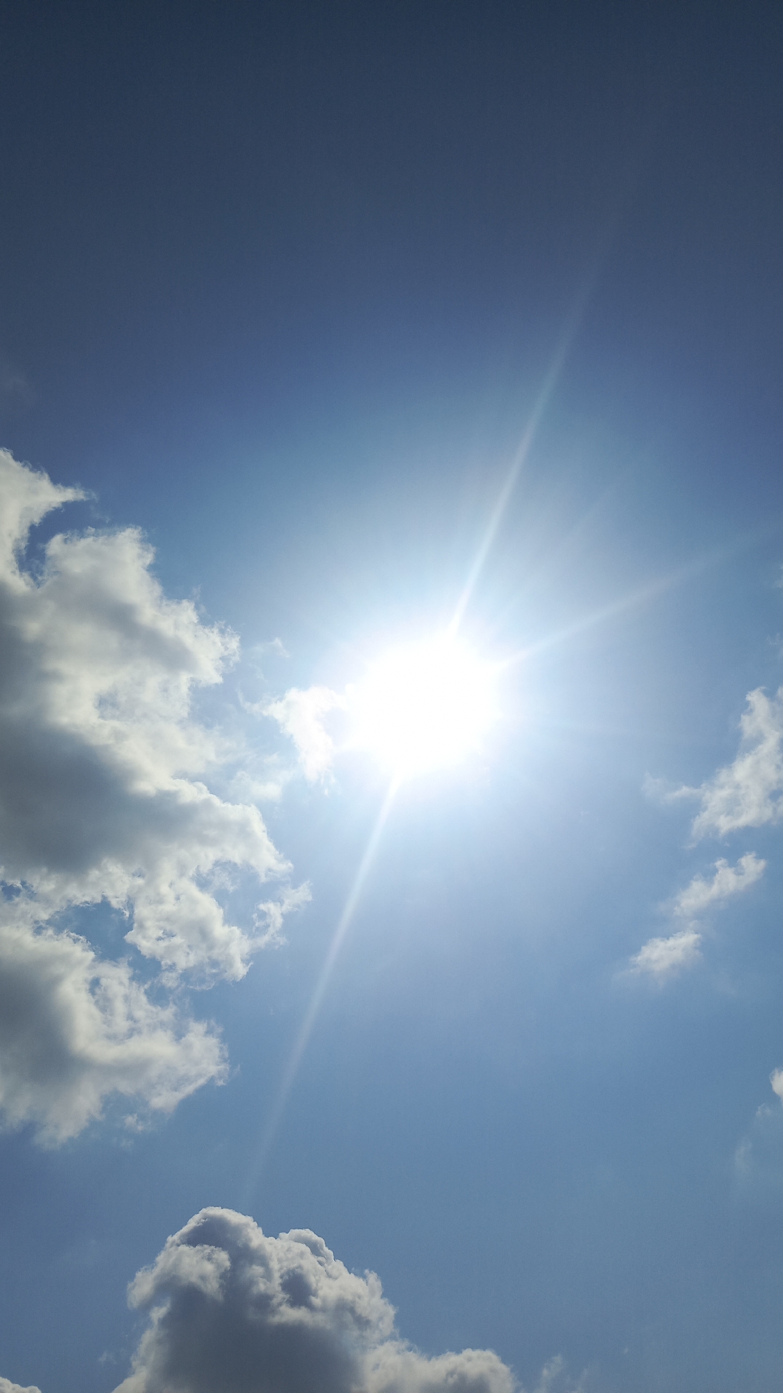 The image is a vertical shot of a bright sun in a blue sky with scattered clouds. The sun is the dominant subject, emitting strong rays that create a lens flare effect. Several fluffy white and gray clouds are visible, particularly on the left side and in the lower portion of the frame. The sky is a clear, vibrant blue. There are no people, buildings, or specific landmarks visible to indicate a location within the city of Ivanovksy, Russia. The scene depicts an outdoor, daytime environment focused on the sky and sunlight.