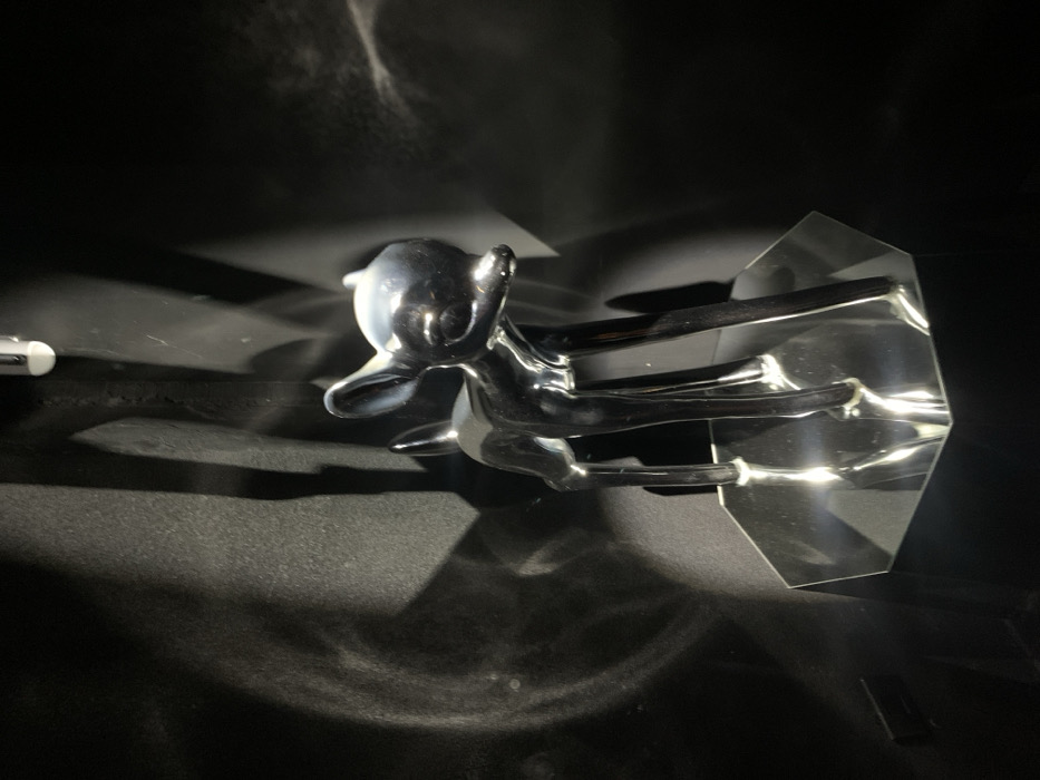 A reflective, chrome-like deer figurine leaps forward, mounted on a geometric, transparent base. The subject sits against a dark, grainy background, catching and reflecting light in a dramatic way. To the side, a partial view of a white object sits. The overall effect is modern and artistic, playing with light and shadow.