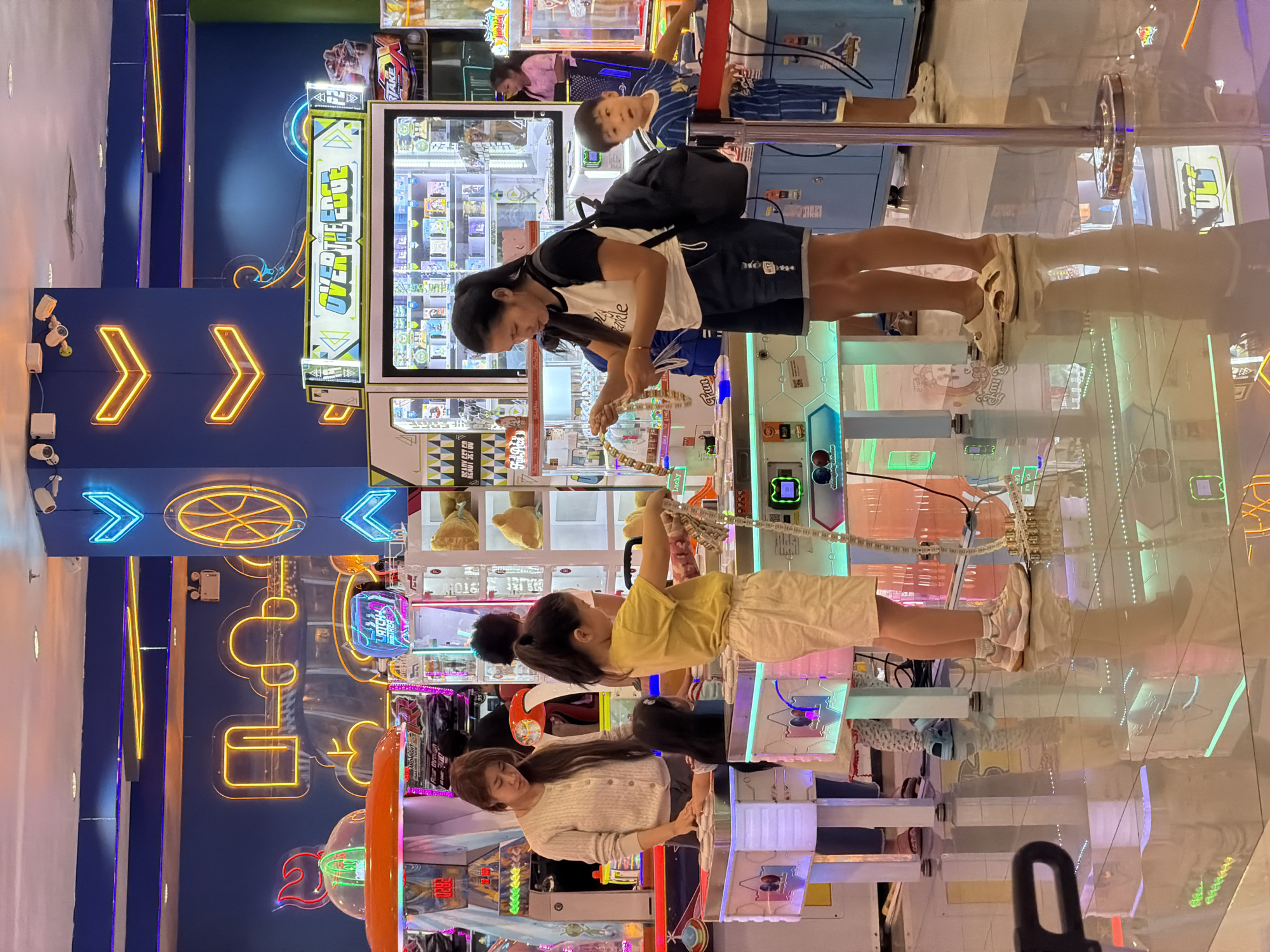 In Chiang Mai City Municipality, Thailand, this image depicts a vibrant arcade scene. Three children and two women are present. One woman, wearing a light-colored cardigan, stands by an arcade game. In the foreground, a young girl in a yellow t-shirt and shorts interacts with an arcade game, while another child, a boy in a blue and white striped shirt, stands nearby. A second woman, wearing a t-shirt and dark shorts, is also engaged with an arcade game, holding what appears to be a prize. The arcade is illuminated by colorful neon lights and features various arcade machines, including prize redemption and skill-based games. The floor is reflective, showing the bright lights and reflections of the arcade.