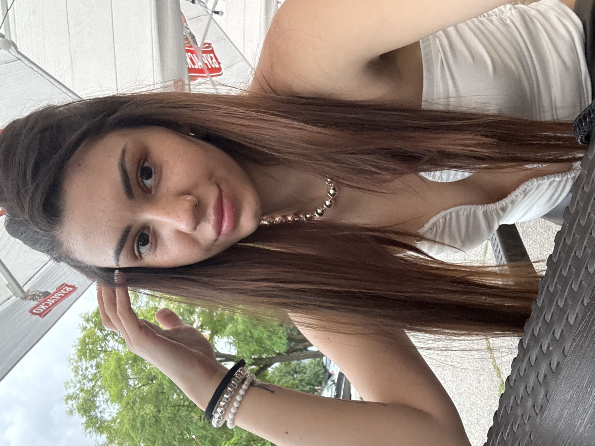 An adult female with long, straight brown hair parted on the side is depicted seated outdoors. Her left hand is raised, with fingers resting near her temple. She has visible freckles on her face, a small hoop piercing in her right nostril, and a ring on her left ring finger. Her left wrist displays a black elastic hair tie, a clear spiral hair tie, and a strand of white beaded bracelets. She wears a white strapless top with a deep V-neckline and a silver-toned, chunky beaded necklace. The foreground includes a dark, textured table surface. The background features a large white commercial umbrella overhead, on which inverted red Cyrillic text "ПИРИНСКО" (PIRINSKO), a Bulgarian beer brand, is visible. Beyond the umbrella, an overcast sky and lush green deciduous trees are discernible. A paved street with partially visible vehicles can be seen through the trees in the distant background. The presence of the "PIRINSKO" branding indicates the location is in Bulgaria, specifically in the city of Sofia.