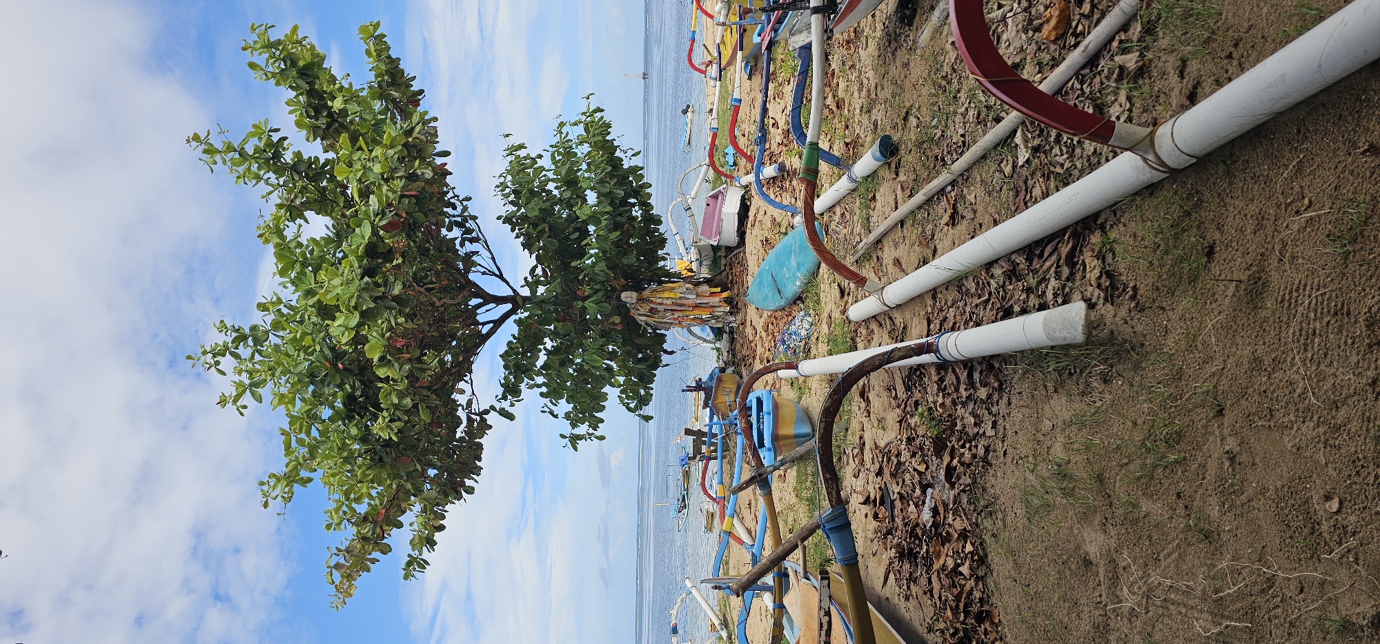 The image depicts a beach scene near Denpasar, Indonesia. Two leafy trees stand at the edge of the beach, adorned with colored streamers or cloths. A collection of outrigger boats and boat parts are scattered on the sand, including colorful bows, bamboo poles, and PVC pipes. The ocean is visible in the background, with several boats on the water. The sky is blue with scattered clouds. The beach itself consists of a mix of sand and dried leaves.