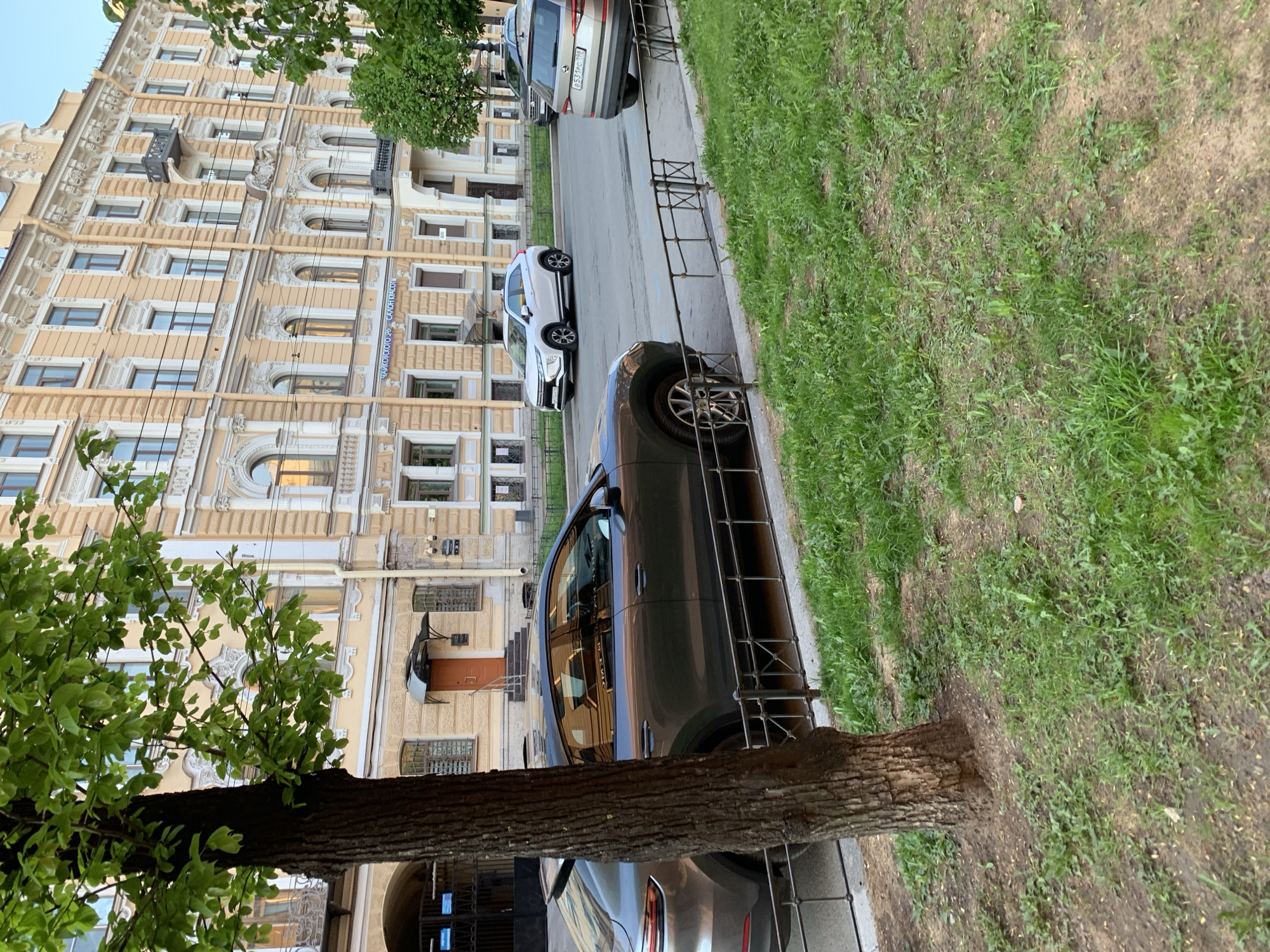 The image depicts a street scene in Saint Petersburg, Russia. An ornate, multi-story building with yellow facade and detailed architectural elements stands prominently on the left side of the frame. Several parked cars are visible along the street. The first is a dark gray car parked parallel to the sidewalk, followed by a white car and a gray SUV. The sidewalk is separated from the street by a black metal railing. A tree trunk is situated on the left. The ground on the right is covered with grass and patches of soil. The image appears to be taken during daylight, with soft shadows indicating even lighting. The license plate of the gray SUV reads: B553 PC 198.