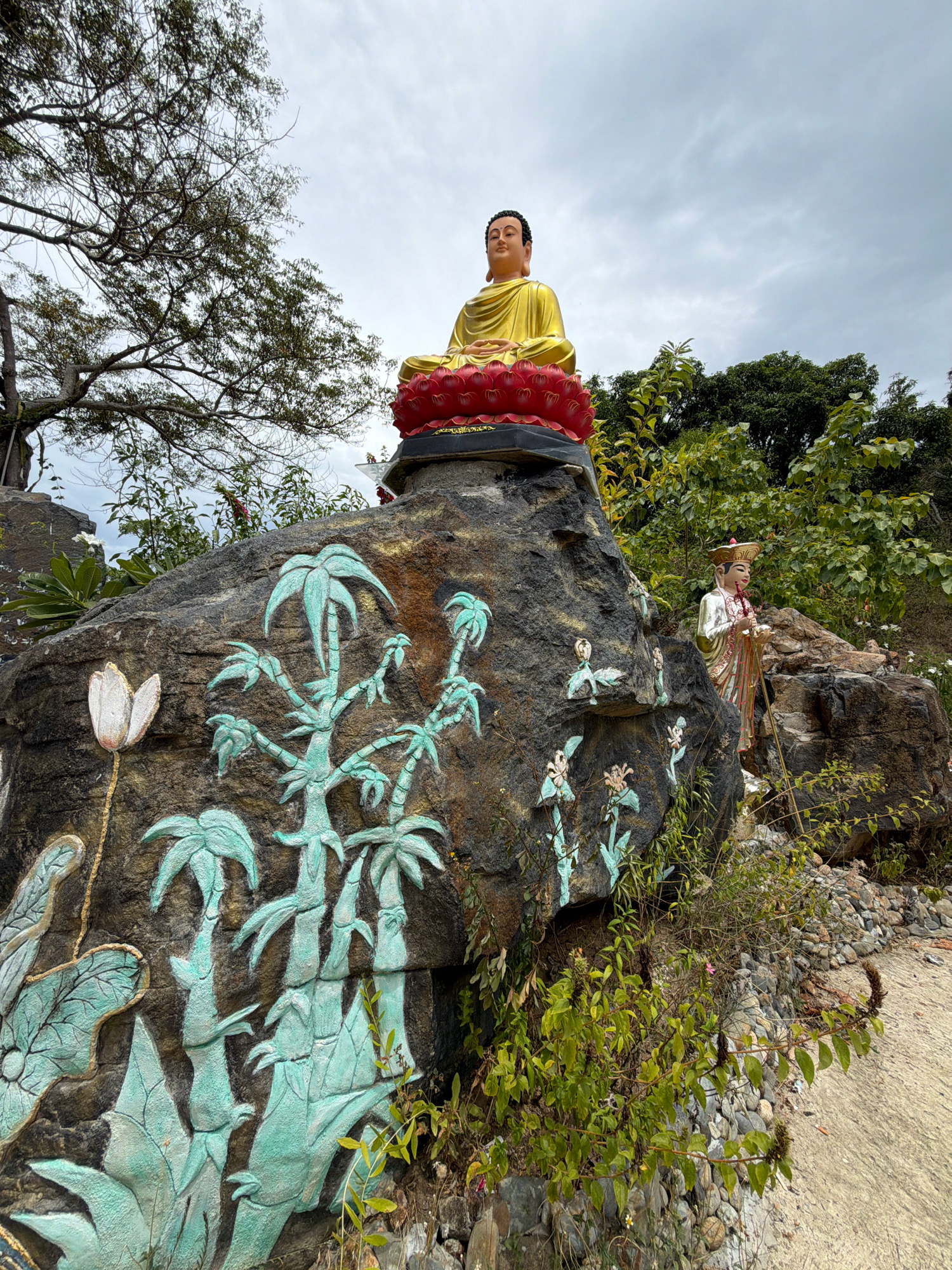 A golden statue of Buddha sits atop a rock formation, dressed in saffron robes. The statue is positioned on a red lotus base. Below the Buddha statue, the rock face is adorned with carved relief sculptures of bamboo and lotus flowers, painted in shades of turquoise and green. To the right of the main rock, a second statue of a figure in traditional attire, holding a staff, stands on a smaller rock. This figure wears a conical hat and a white and gold robe. The surrounding environment is lush with greenery, including trees and various plants, some of which are growing around the rock formations. The sky above is overcast. The scene appears to be an outdoor religious site, possibly a temple or garden, located in Nha Trang, Vietnam, characterized by its natural elements and religious iconography.