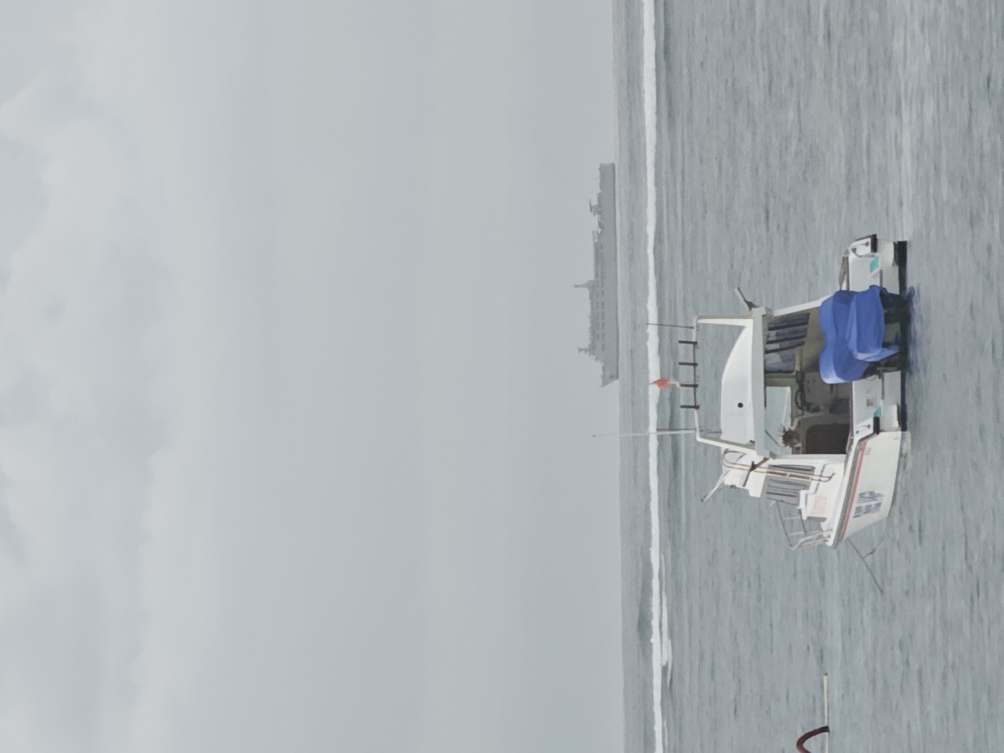 The image captures a serene ocean scene under an overcast sky. In the foreground, a white motorboat with blue tarps covering part of its stern is moored in calm waters. The boat has a cabin and a railing. The stern of the boat displays red and white stripes along its waterline, and text that appears to read "TIME UP". In the middle ground, a larger vessel, likely a ferry or cargo ship, is visible on the horizon, silhouetted against the hazy sky. The sea has gentle waves, with white surf lines indicating shallow areas or a break in the water. A red flag is attached to a pole on the motorboat, and a white pole is visible to the right of the motorboat. The overall impression is one of a cloudy day at sea, with two vessels of different sizes present. The location is likely near the coast of Denpasar, Indonesia, given the presence of similar boats and the general seascape.
