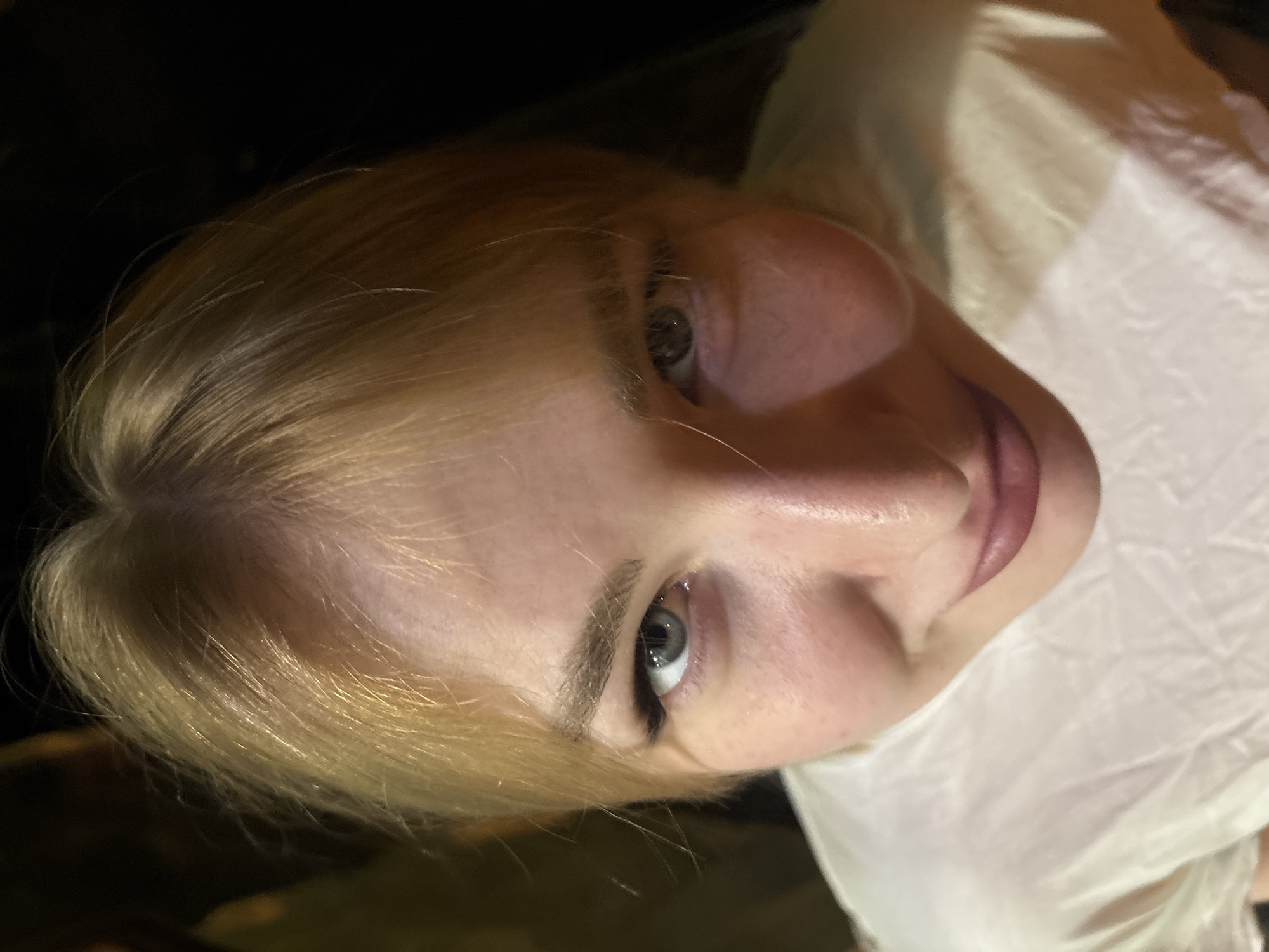 The image shows a close-up, high-angle shot of a young Caucasian woman with blonde hair and light-colored eyes. She is wearing a white shirt. Her facial features are prominent, including her eyebrows, eyes, nose, and lips. The background is dark and out of focus, obscuring any details of the environment. There are no discernible indications of the city of Khimki, Russia, or any specific location within it. The image appears to be a portrait taken in low light conditions.