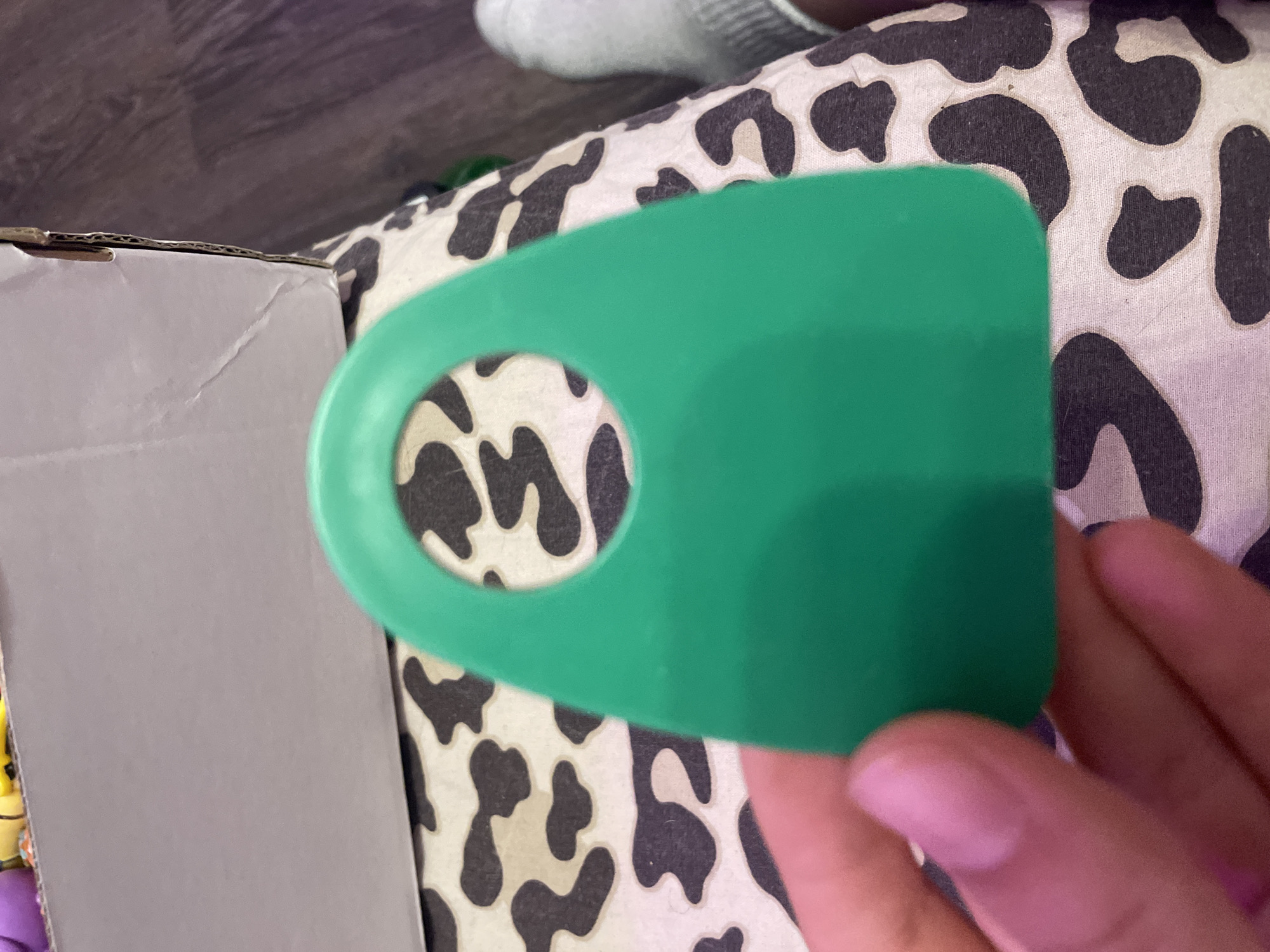 In the city of Vaganovo, Russia, a person's hand with visible fingernails is holding a bright green, plastic object with a rounded top and a circular cutout in the center. The object is positioned over a surface with a black and white cow-print pattern. In the background, a cardboard box is visible, with the edge of a colored item peeking out from inside. A person's foot wearing a gray sock is also visible on the right side of the frame. The overall setting appears to be indoors, with a wooden floor visible in the far background.