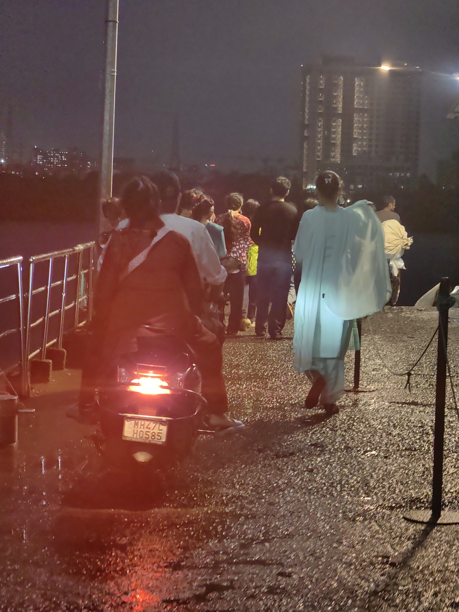 In Gorai, India, under a dimly lit, possibly foggy or rainy sky, a group of people are gathered on a paved walkway next to a metal railing. A motorcycle with its brake lights illuminated is in the foreground, displaying the license plate "MH47C H0585". Several individuals are visible, including a person on the motorcycle wearing a red garment and another person in front of them wearing white. Further back, more people are seen, some in casual attire. In the background, a tall, modern building with numerous lit windows is visible, suggesting an urban or suburban setting. The ground is wet, indicating recent rain.