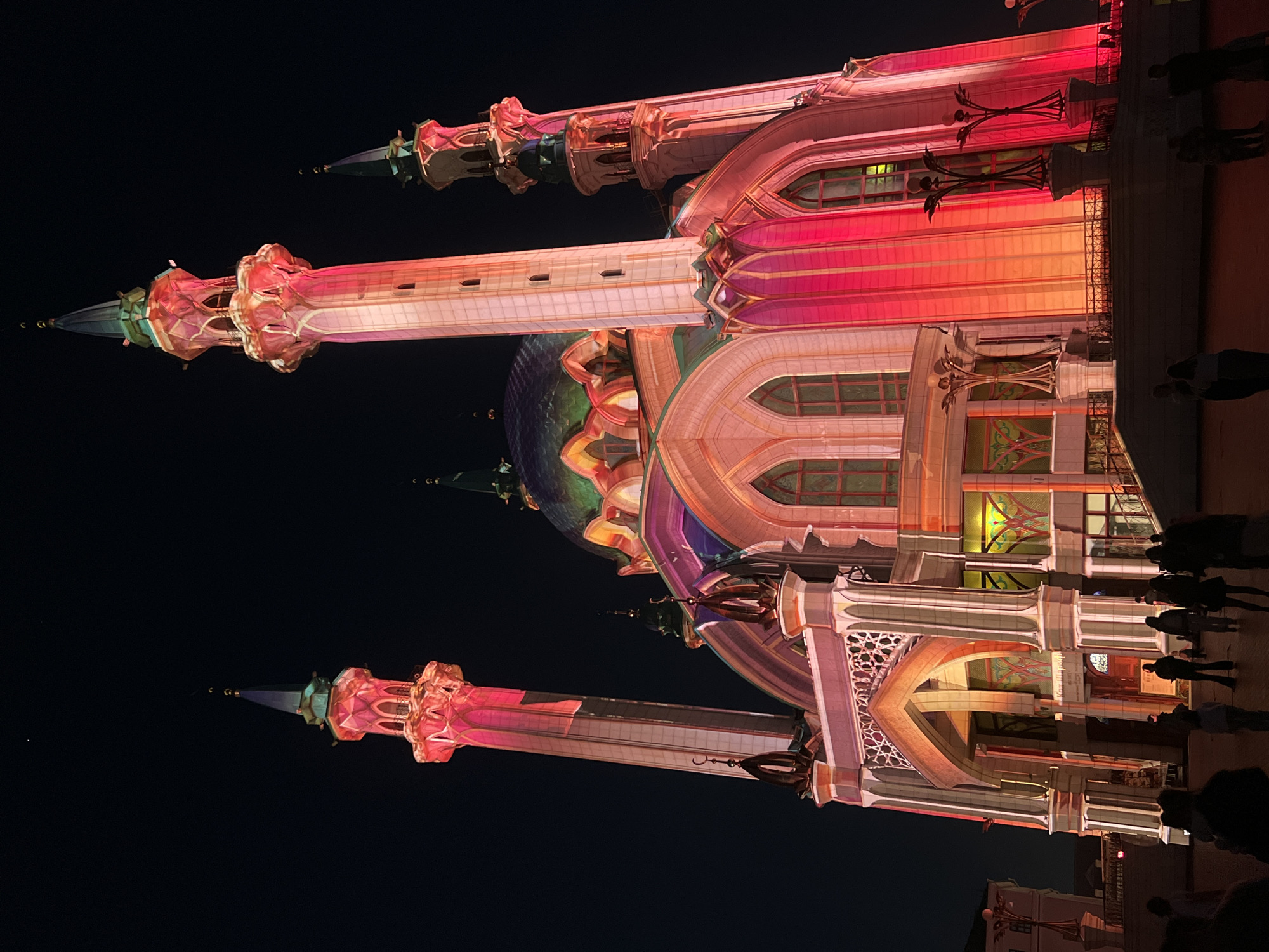 The image shows the Kul Sharif Mosque in Kazan, Russia, at night. The mosque is illuminated with a colorful light display, featuring shades of pink, orange, and white. Three minarets are visible, and people are walking on the ground near the building. The night sky is dark.
