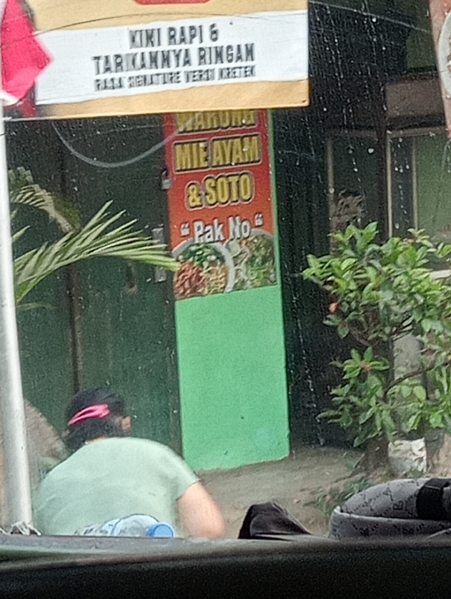 The image captures a street view in Magelang, Indonesia, featuring a food stall signage and a person. The prominent sign, colored red and green, displays text that reads "WARUNG MIE AYAM & SOTO PAK NO". Above this, another sign in white and yellow states "KIKI RAPI & TARIKANNYA RINGAN RASA SIGNATURE VERSI KRETEK". A person, seen from behind, is wearing a light green t-shirt and a pink headband, sitting in what appears to be a vehicle. A plastic water bottle is visible in the foreground. The environment suggests an outdoor setting, with greenery from a plant on the right side of the frame. The weather appears to be rainy or recently rained, as evidenced by streaks on the glass through which the photo is taken and possible water droplets on the background.