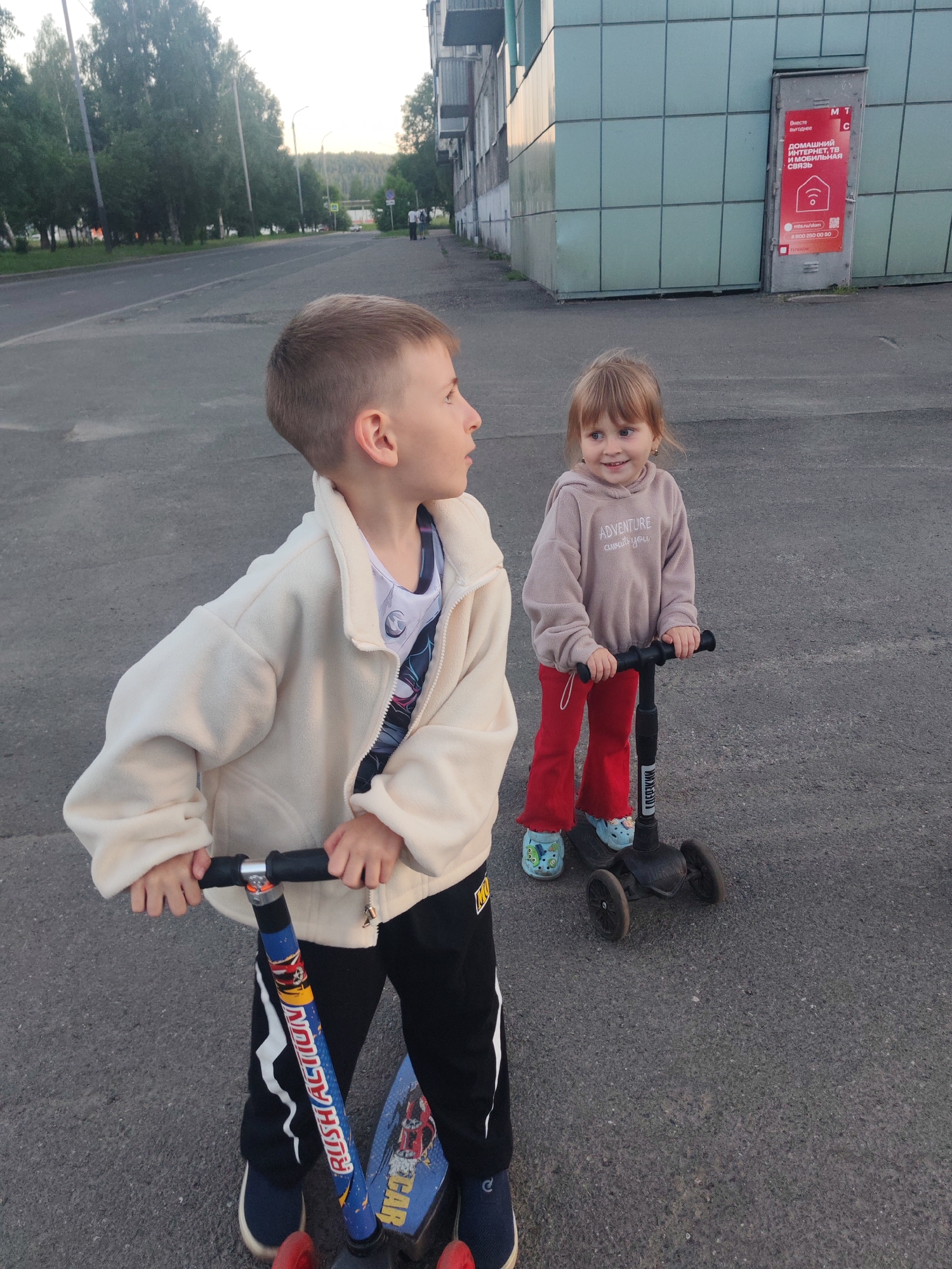 A young boy and girl are outdoors on a paved surface, likely a sidewalk or street, in front of a modern building. The boy, on the left, is wearing a cream-colored jacket over a graphic t-shirt and black pants. He is holding the handlebars of a scooter with a blue and red graphic design on its stem. The girl, on the right, is wearing a beige hoodie with the word "ADVENTURE" printed on it, red pants, and blue clogs with cartoon characters. She is also on a scooter, which is black. The building behind them has a facade of light blue, grid-like panels and a gray metallic door with a red advertisement. The background shows a street with trees lining one side and a distant view of a wooded area under a pale sky. The image appears to be taken in Mezhdurechensk, Russia, judging by the building's architecture and the general urban setting.